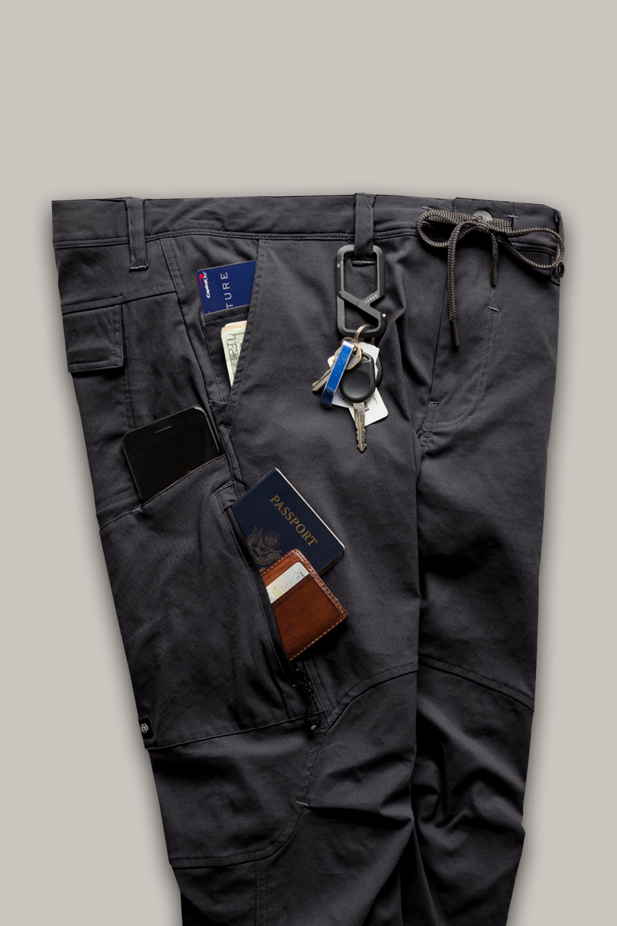 The 686 Men's Anything Cargo Pant - Relaxed Fit in dark gray, shown against a plain light background, features 13 pockets holding a phone, passport, wallet, cash, and keys attached to a carabiner.