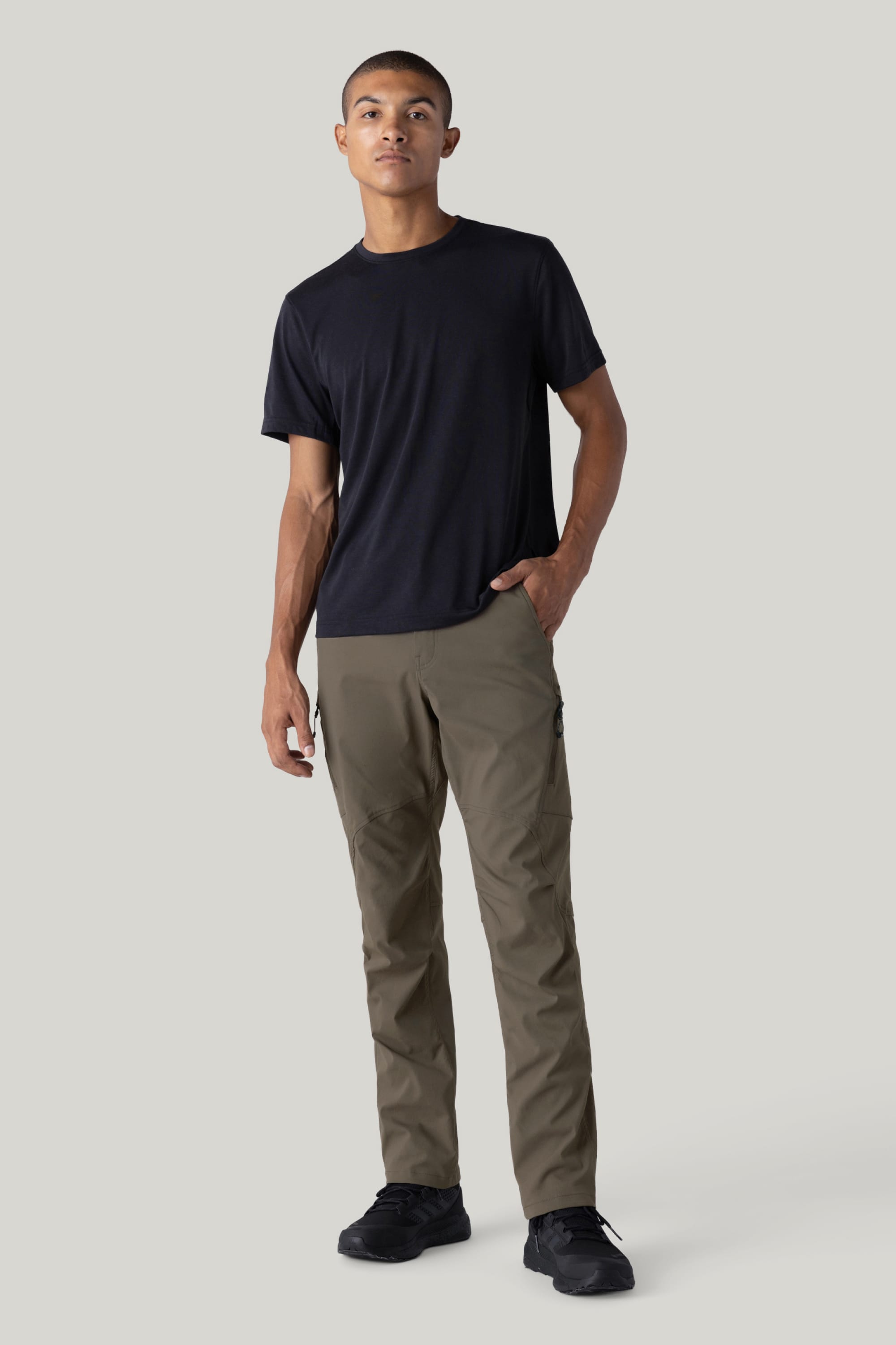A person stands against a plain light gray background wearing a black t-shirt, the 686 Men's Anything Cargo Pant - Relaxed Fit in olive green with a 13-pocket design, and black shoes, one hand in their pocket and the other relaxed at their side.