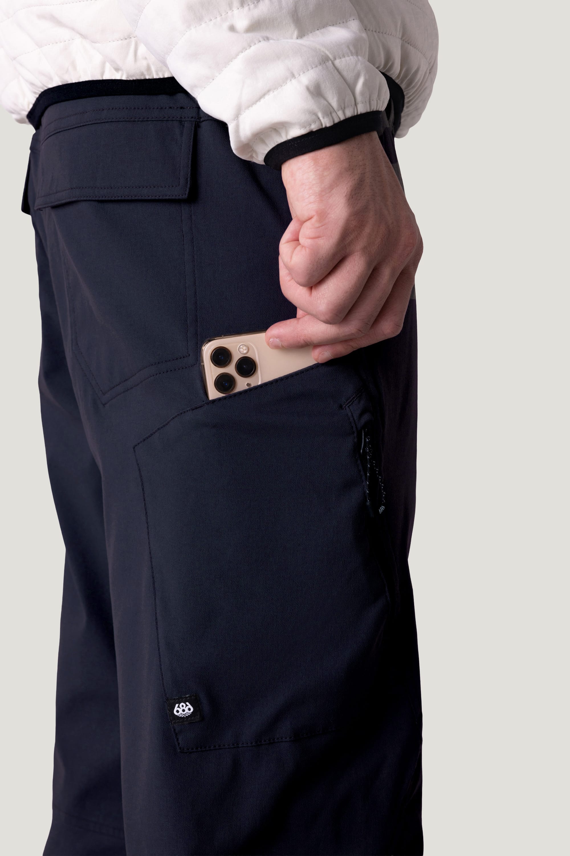 A person wearing 686 Men's Anything Cargo Pant - Relaxed Fit in dark color and a white jacket places a triple-lens smartphone into the side zippered pocket of their 686 cargo pants.