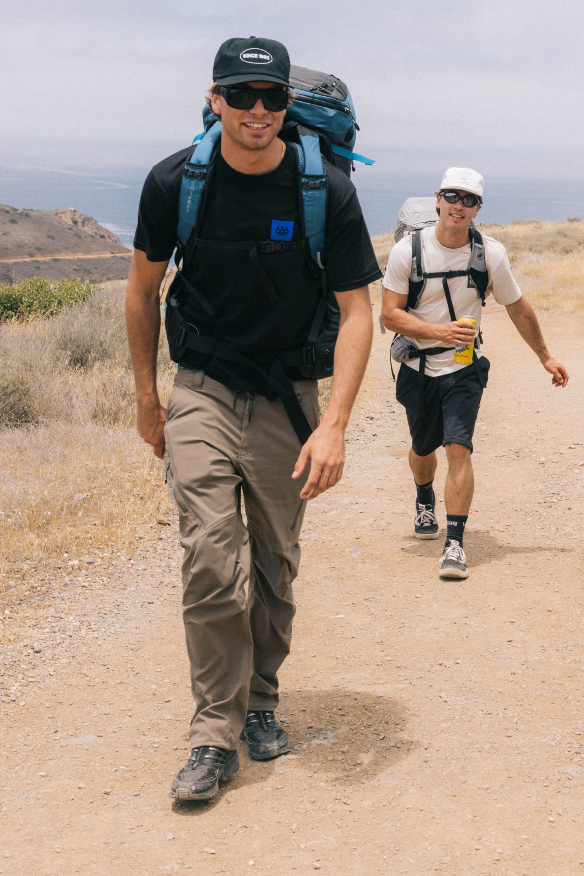 Two men in hats, sunglasses, and backpacks hike a dry coastal trail. The man in front smiles as the other carries a drink—both wearing 686 Men's Anything Cargo Pant - Relaxed Fit, designed by 686 for comfort and easy movement.