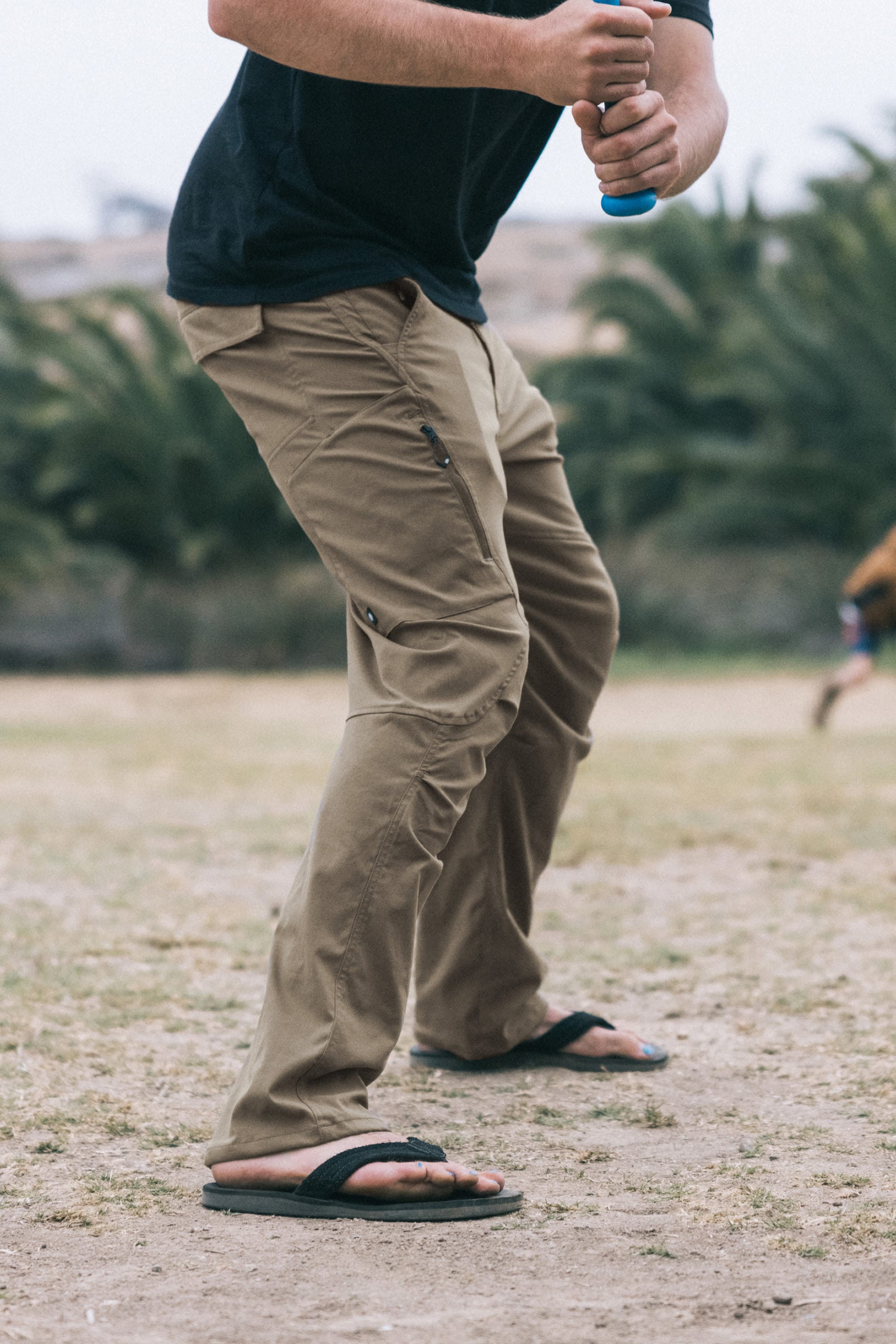A person in 686 Men's Anything Cargo Pant - Relaxed Fit (tan), a black shirt, and black flip-flops stands outdoors with a blue and white bat, ready to swing. The grassy background has blurred trees and someone running.