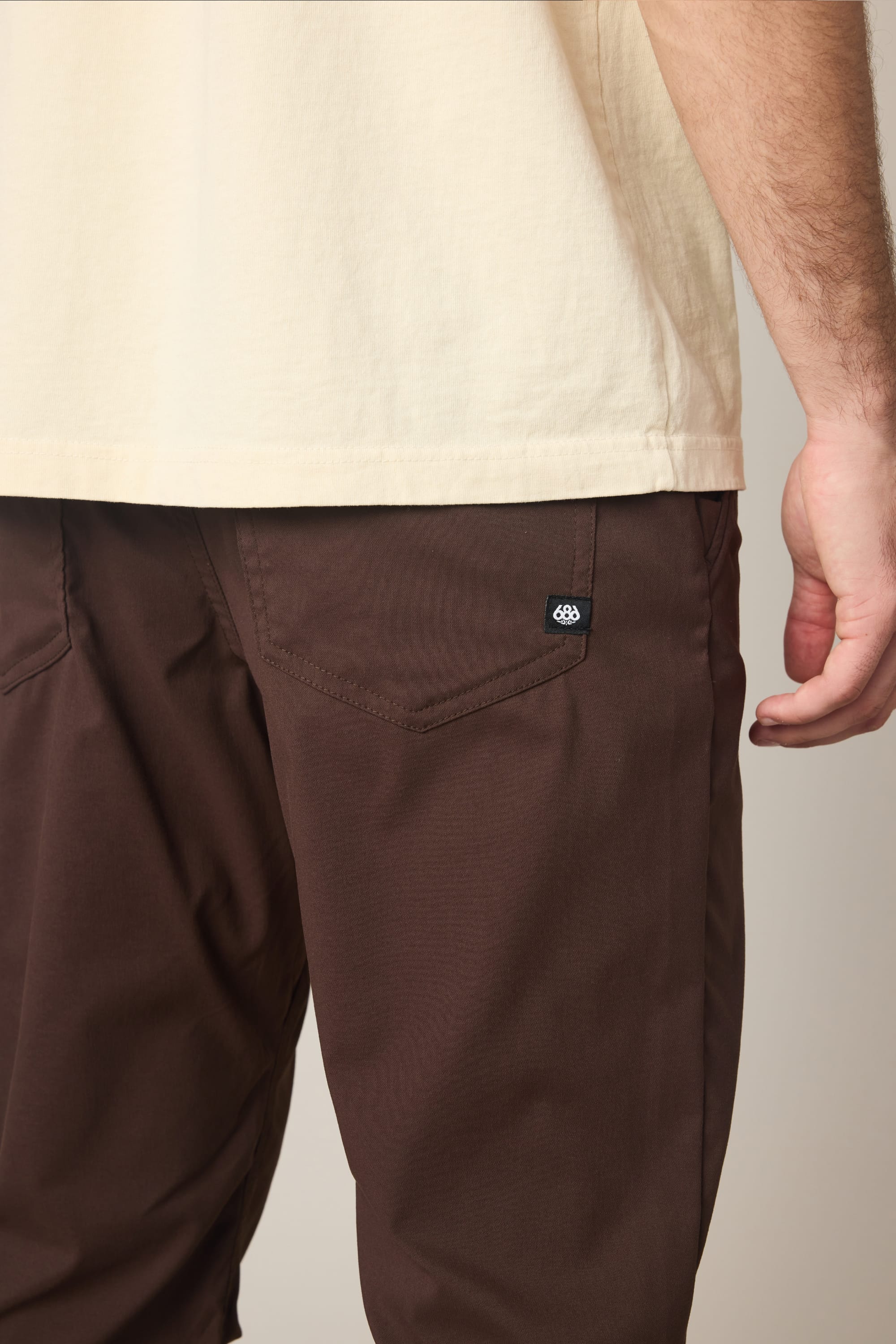 A person is shown from the waist down wearing a cream-colored shirt and 686 Men's Everywhere® Pant - Relaxed Fit in brown, with their right hand relaxed at their side. A small black 686 logo tag is visible on the right back pocket of the breathable pants.