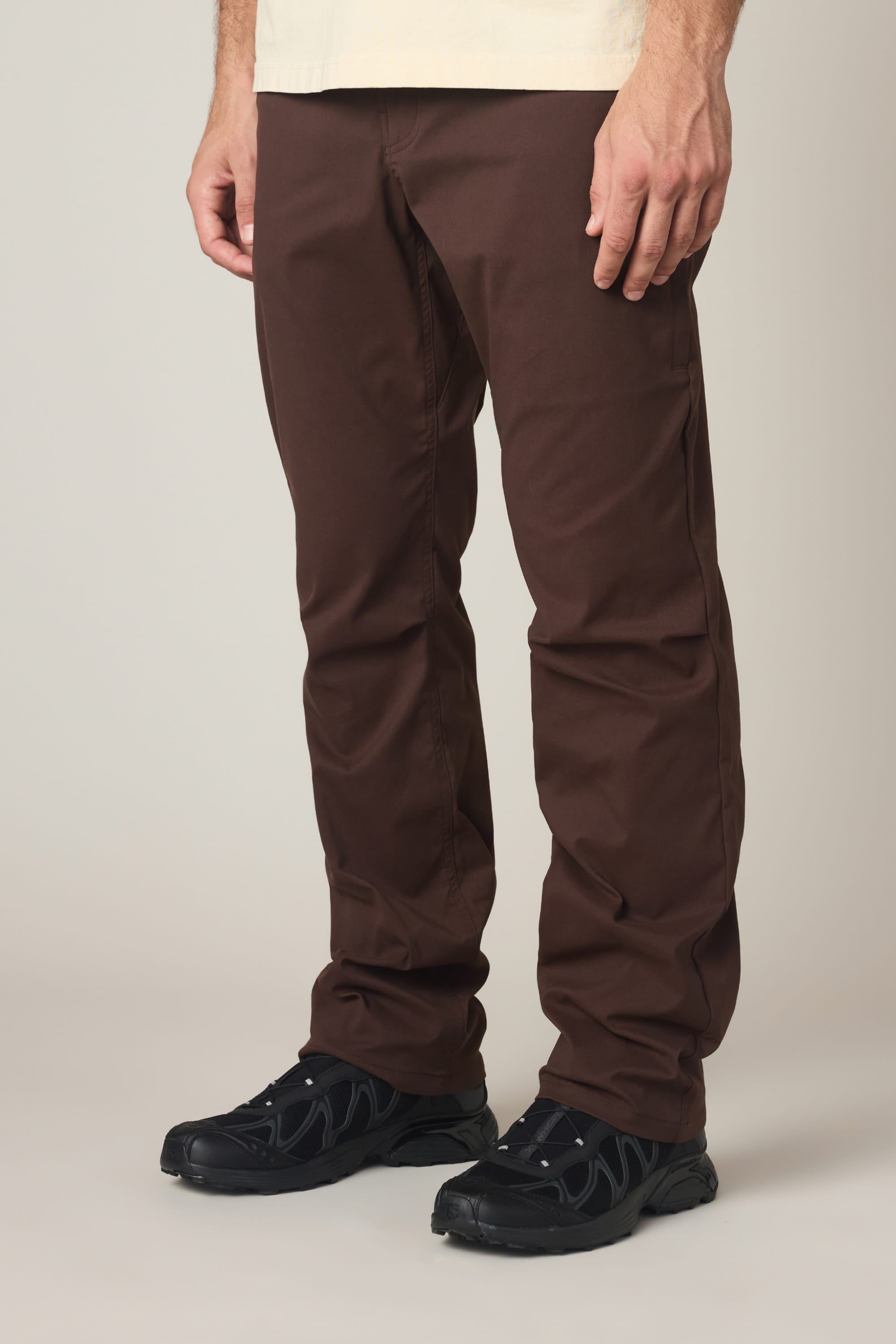 A person is shown from the waist down wearing 686 Men's Everywhere® Pant - Relaxed Fit in brown, paired with black sneakers, standing against a plain light background.