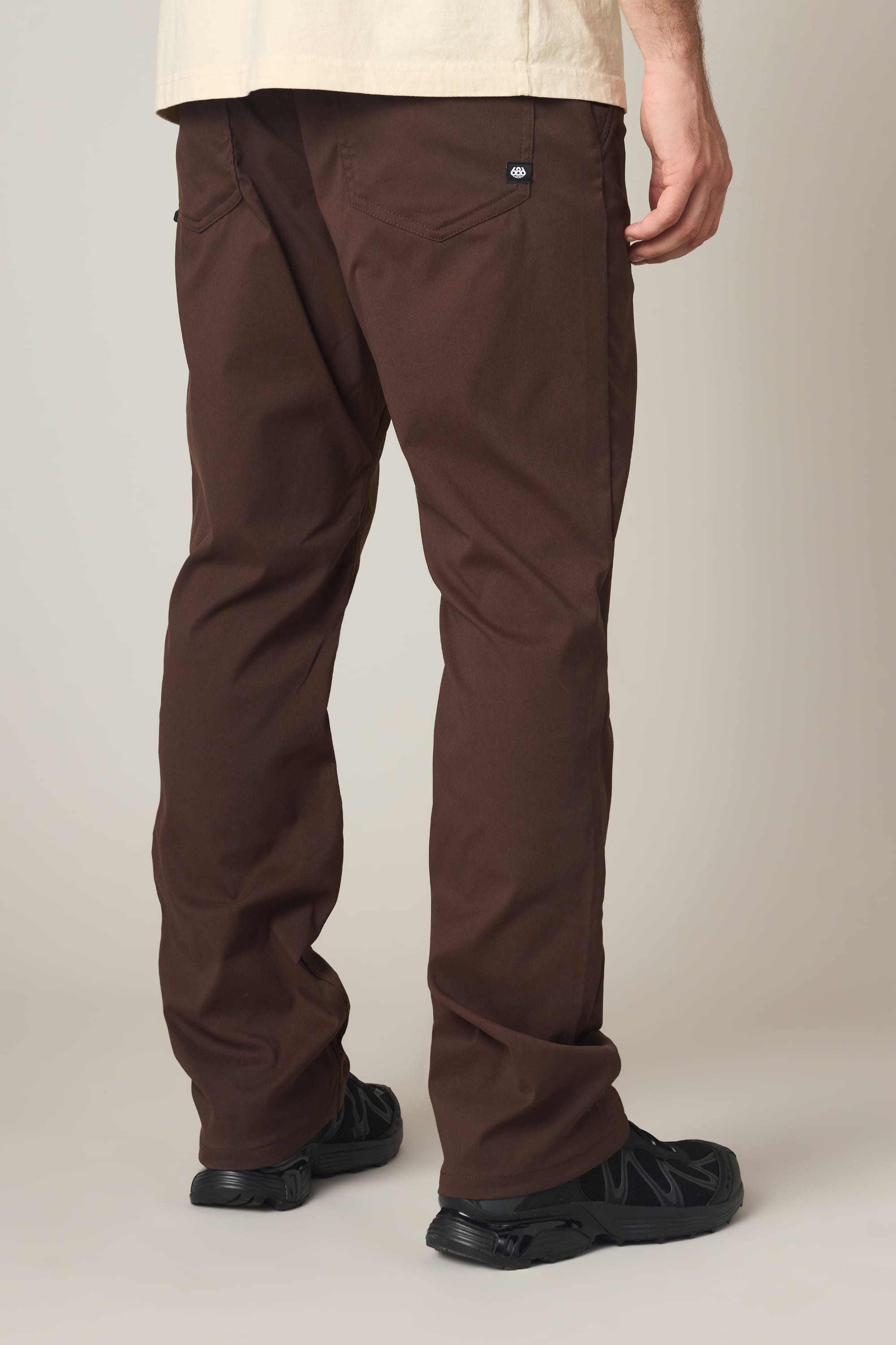 A person wearing the 686 Men's Everywhere® Pant - Relaxed Fit in brown, a beige shirt, and black shoes is standing with their back to the camera.