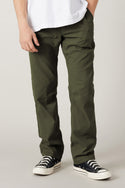 A person models the 686 Men's Everywhere® Pant - Relaxed Fit in olive green with a white t-shirt and black low-top sneakers with white soles, standing against a plain light background. Their face is not visible.