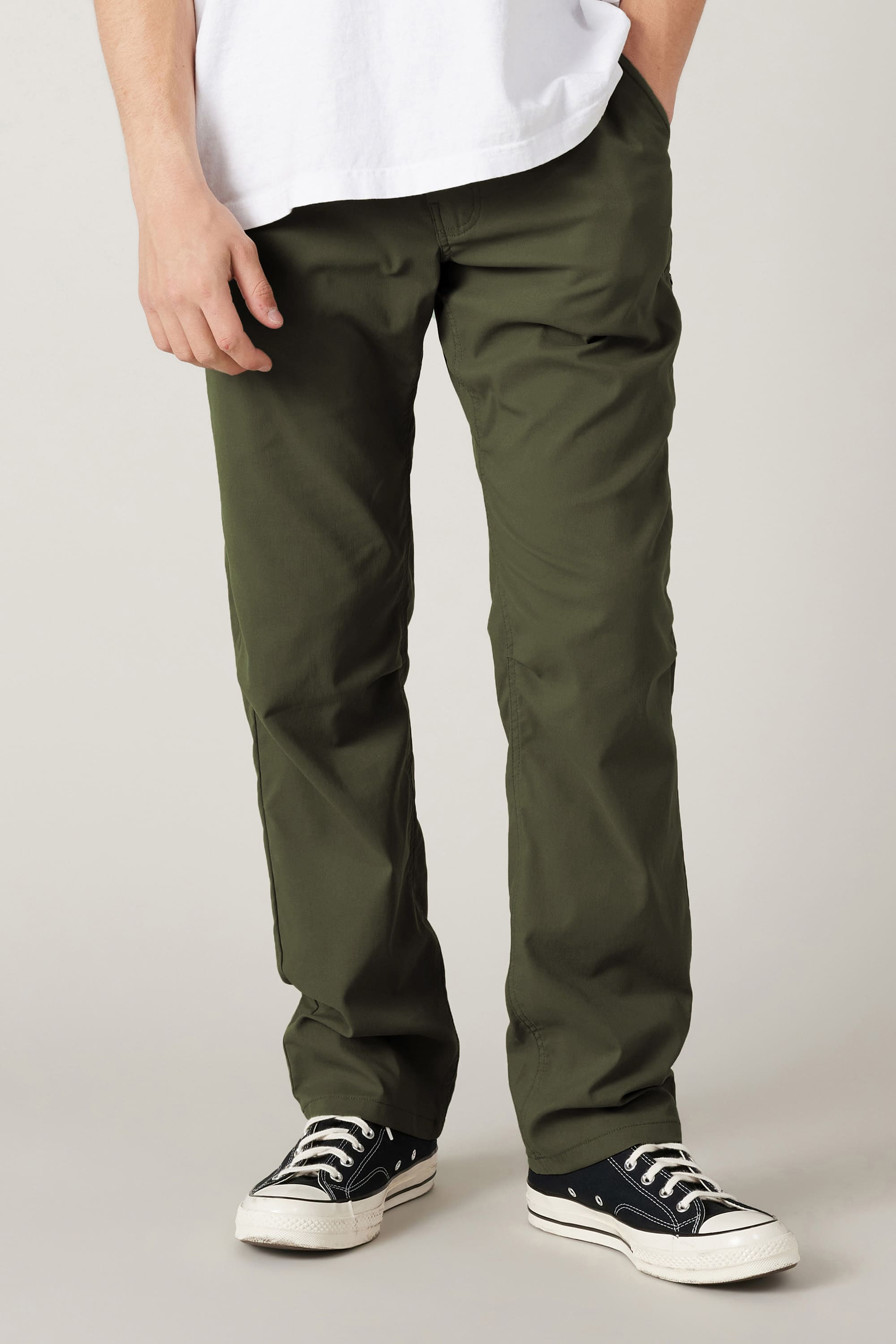 A person models the 686 Men's Everywhere® Pant - Relaxed Fit in olive green with a white t-shirt and black low-top sneakers with white soles, standing against a plain light background. Their face is not visible.