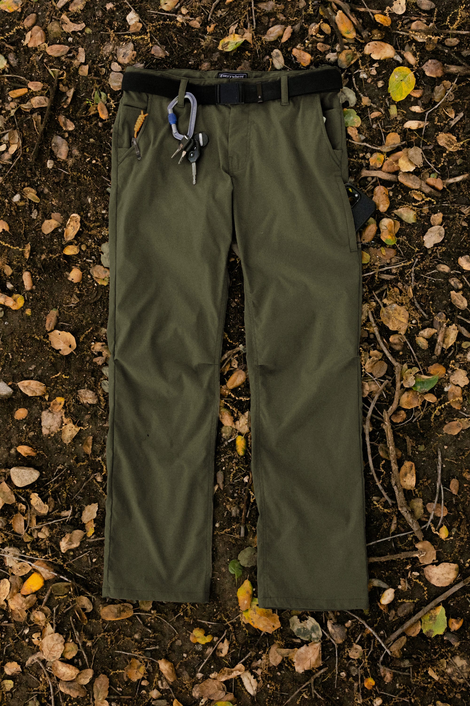 The 686 Men's Everywhere® Pant - Relaxed Fit in olive green, featuring a black belt and climbing gear on the front belt loop, is displayed flat on soil scattered with yellow and brown leaves.
