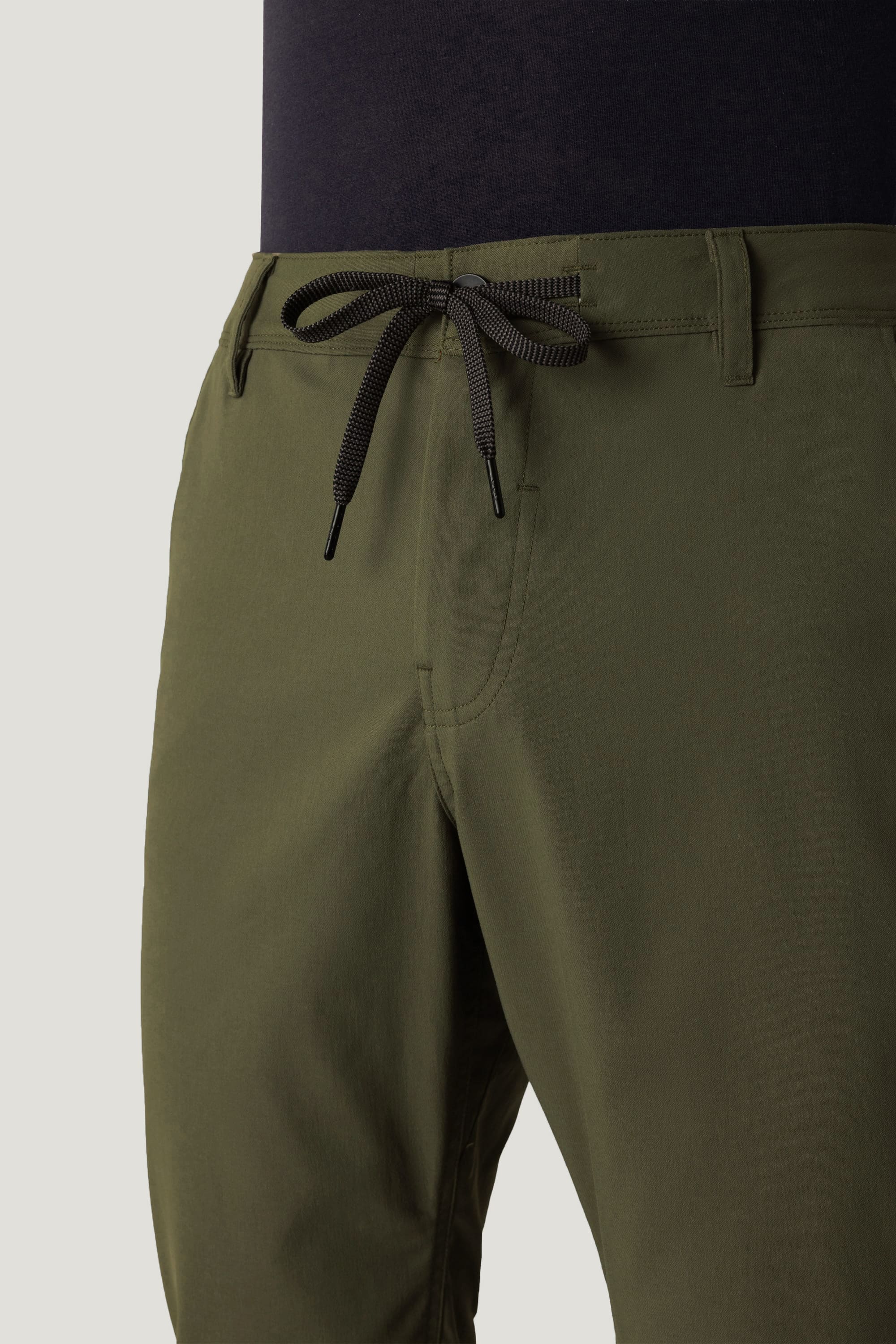 A close-up of a person wearing the 686 Men's Everywhere® Pant - Relaxed Fit in olive green with a black drawstring waist, paired with a dark shirt tucked in.