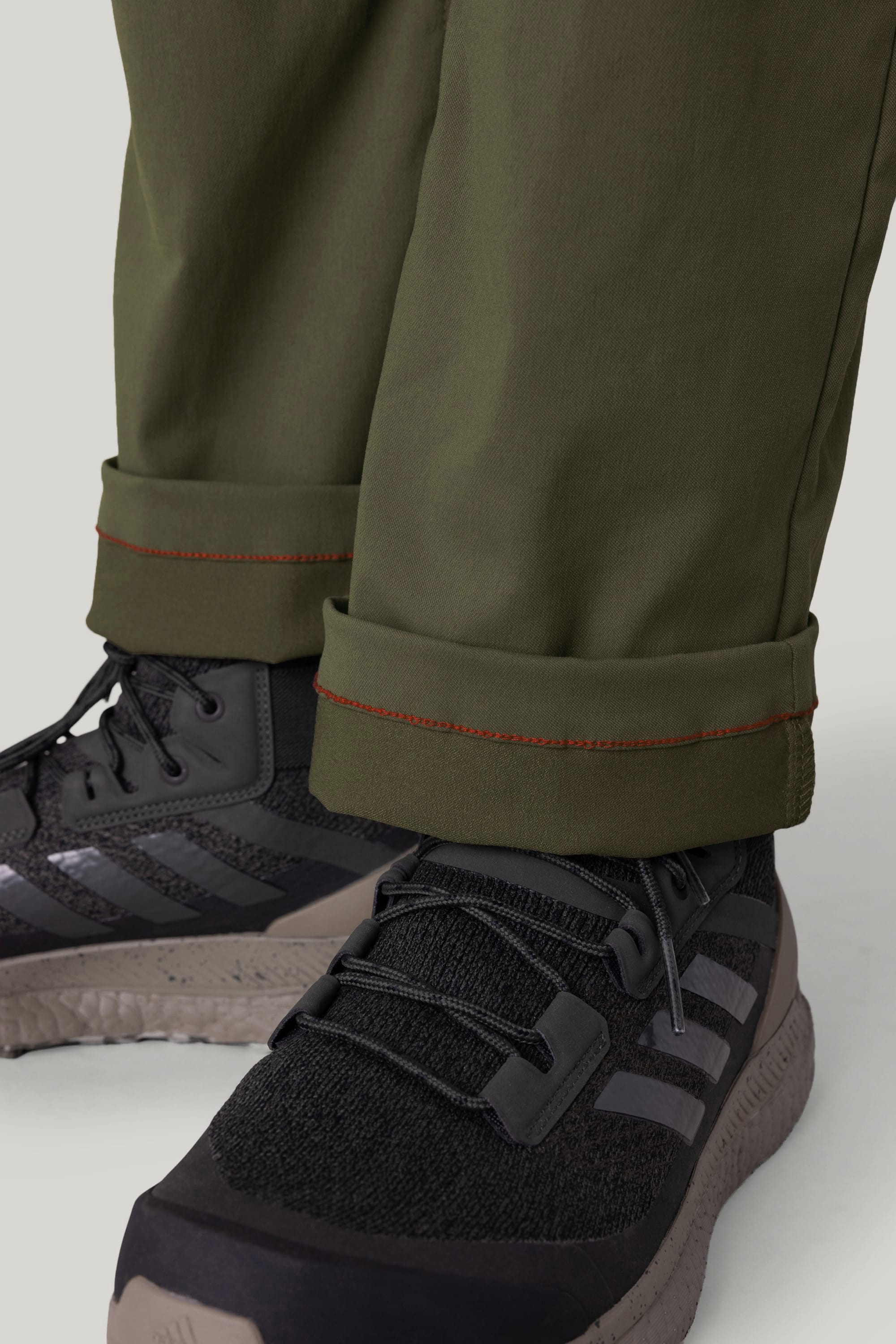 Close-up of a person wearing the 686 Men's Everywhere® Pant - Relaxed Fit in dark green with rolled cuffs and orange stitching, paired with black athletic shoes featuring gray stripes and thick soles.