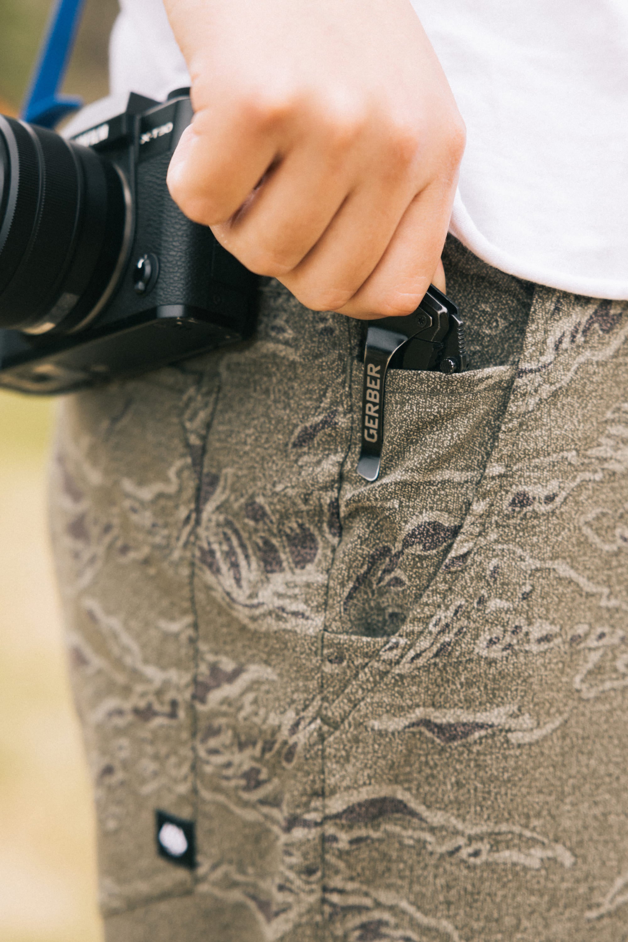 A person wearing 686 Men's Everywhere Pant - Relaxed Fit in a camouflage pattern holds a Gerber pocket knife clipped to their pocket and carries a black camera around their neck.