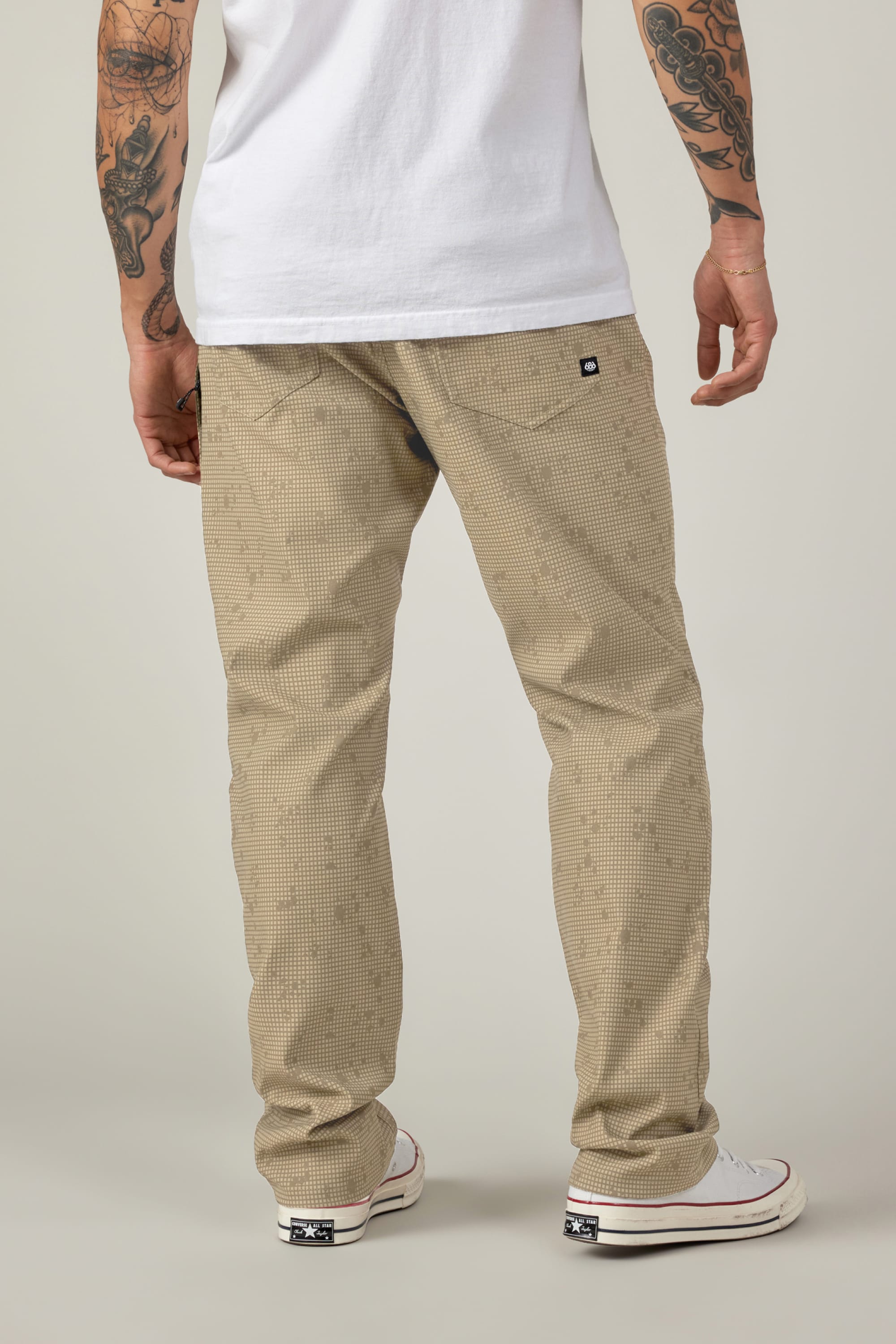 A person is shown from the back wearing white sneakers, a white t-shirt, and 686 Men's Everywhere® Pant - Relaxed Fit by 686 in beige check with a small black logo on the pocket; both arms are tattooed.