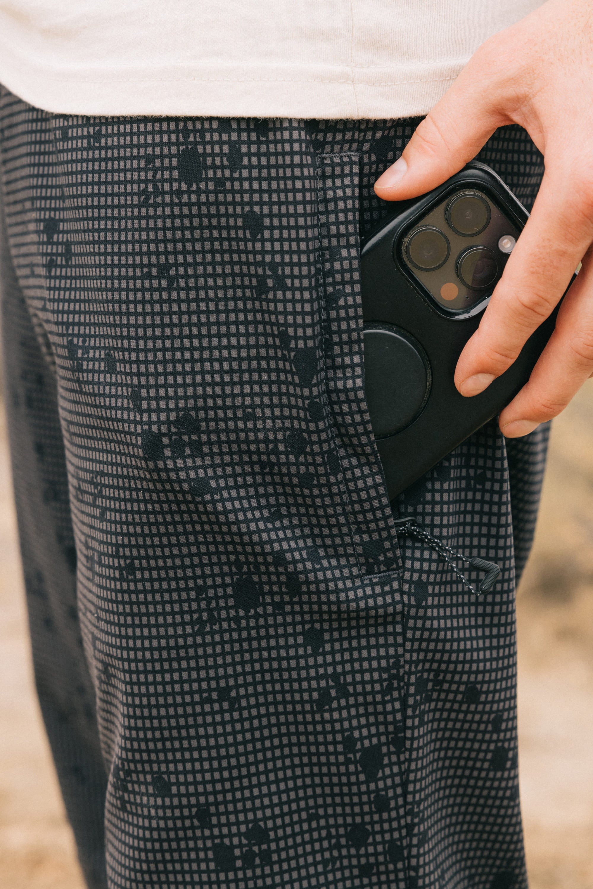 A hand slips a black smartphone into the side pocket of 686 Men's Everywhere Pant - Relaxed Fit. The pants are dark and patterned; only the person's torso and hand are visible against a blurred background.