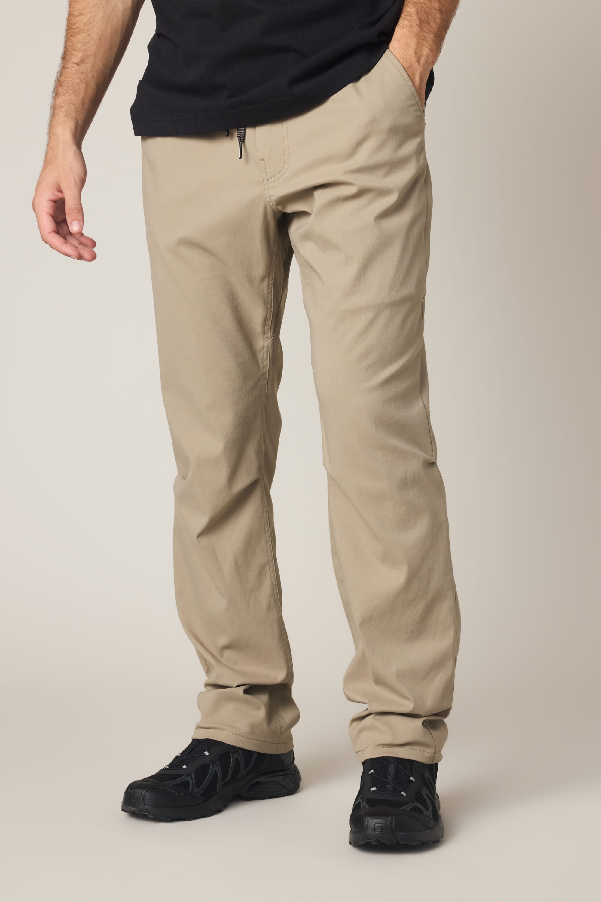 A person wearing the 686 Men's Everywhere® Pant - Relaxed Fit in beige, paired with a black t-shirt and black shoes, stands with one hand in their pocket against a neutral background.