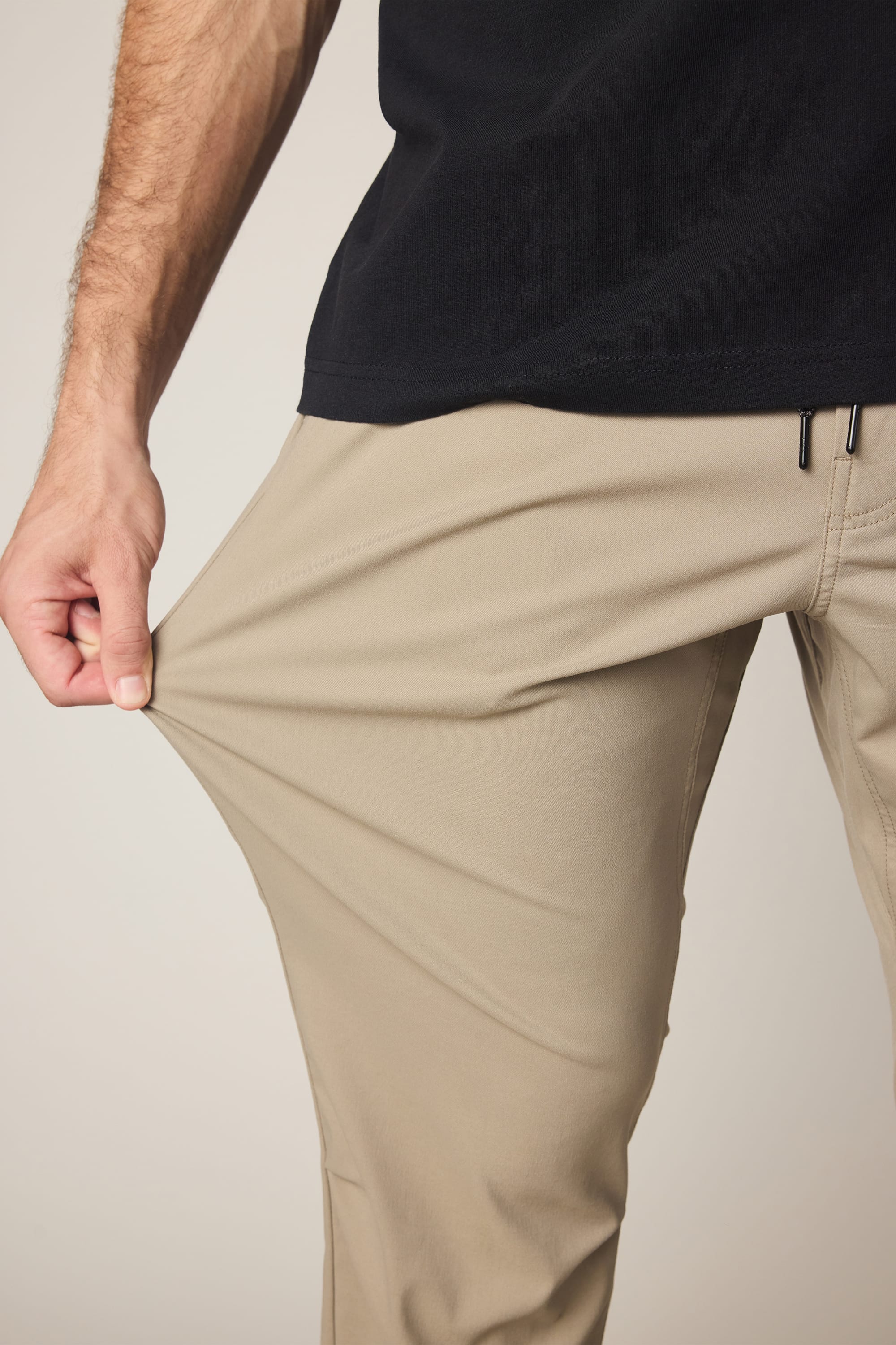 A person wearing 686 Men's Everywhere® Pant - Relaxed Fit in tan and a black shirt stretches the fabric of their pants outward with one hand, demonstrating its flexibility.