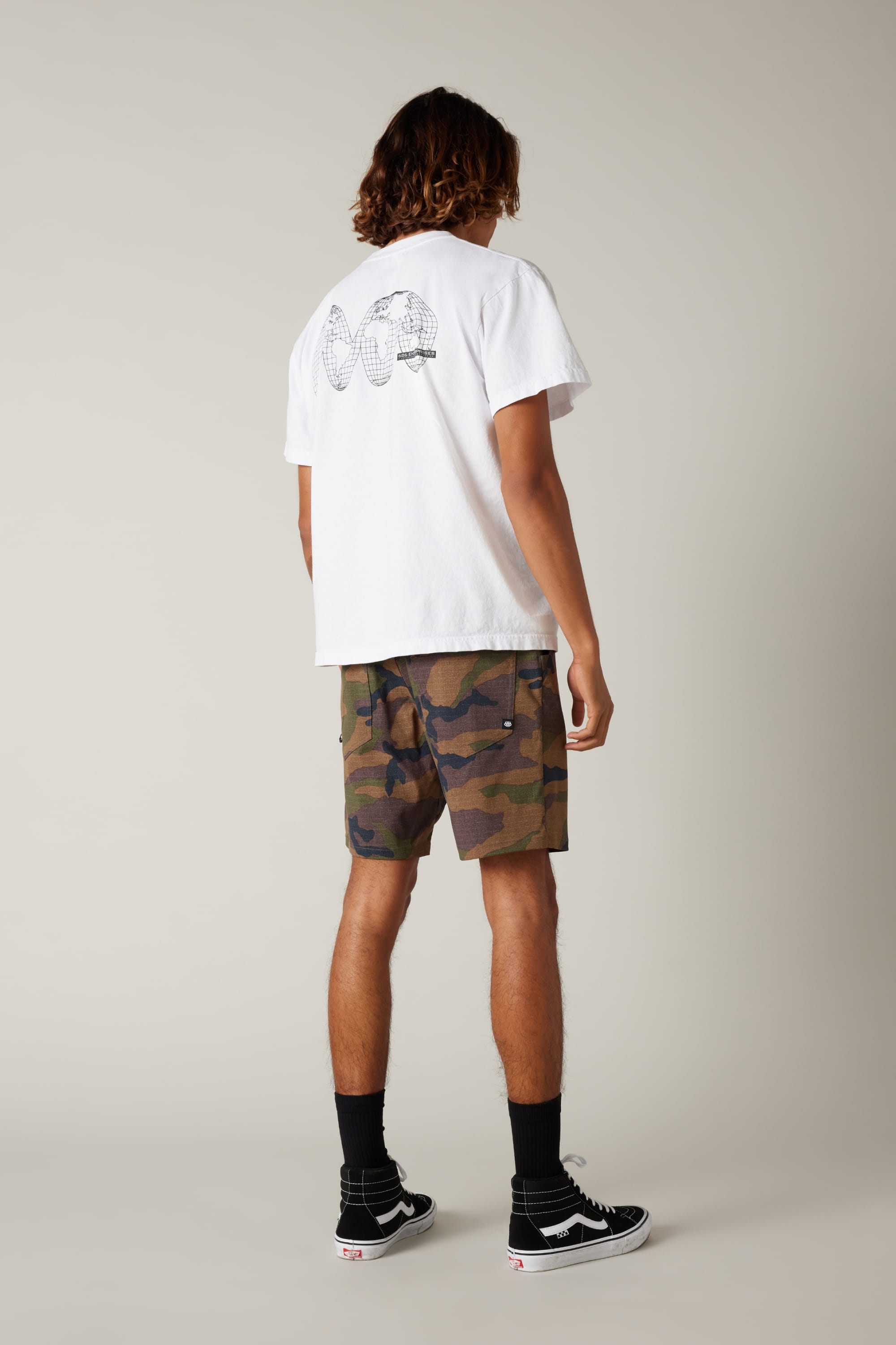 A person faces away, wearing a white graphic tee, 686 Men's Everywhere® Hybrid Short by 686 in a camo print, black crew socks, and black Vans sneakers against a neutral background.