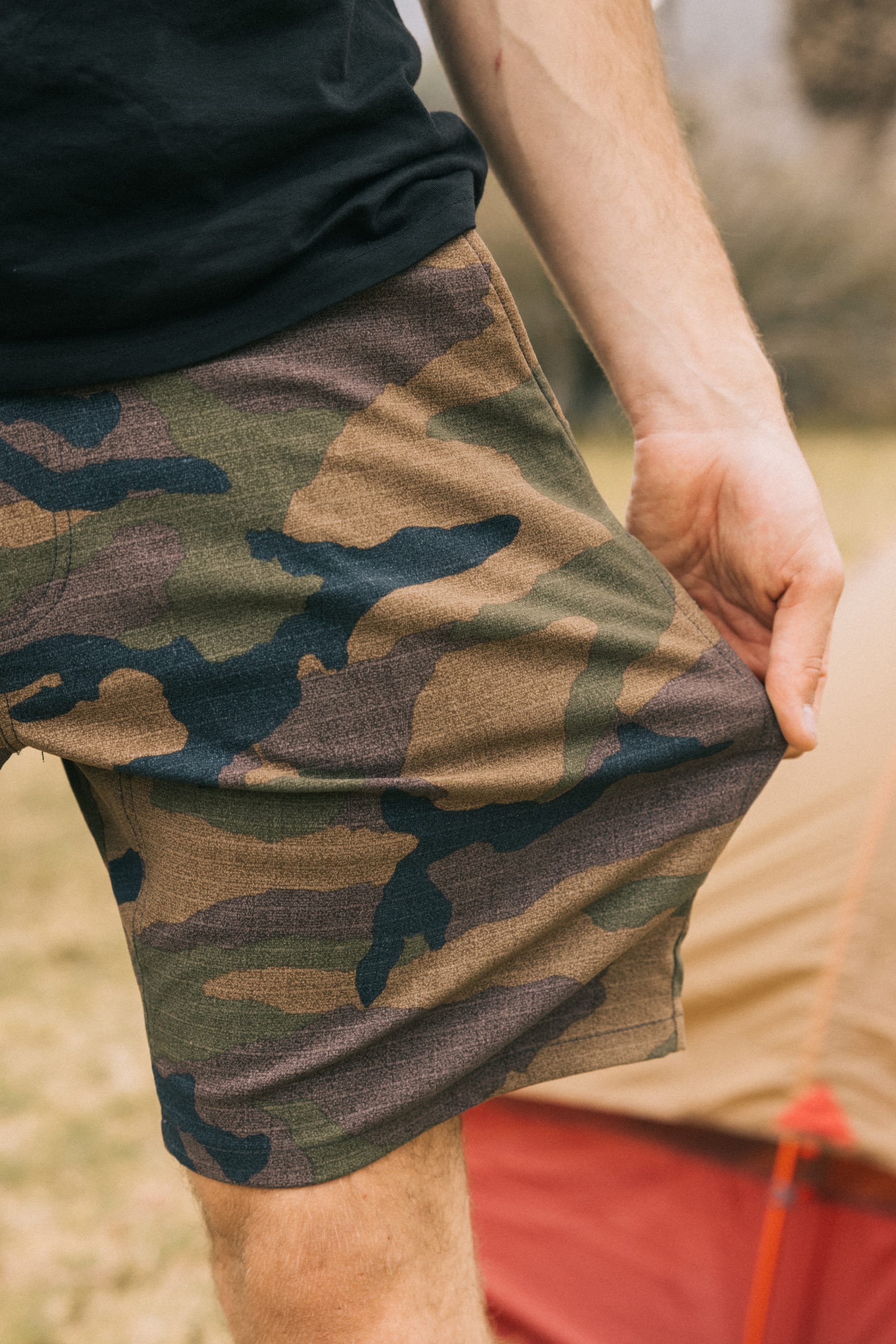 A person wearing 686 Men's Everywhere® Hybrid Short and a black shirt stands outdoors, holding the hem of their shorts. A tan and red tent is visible in the blurred background.