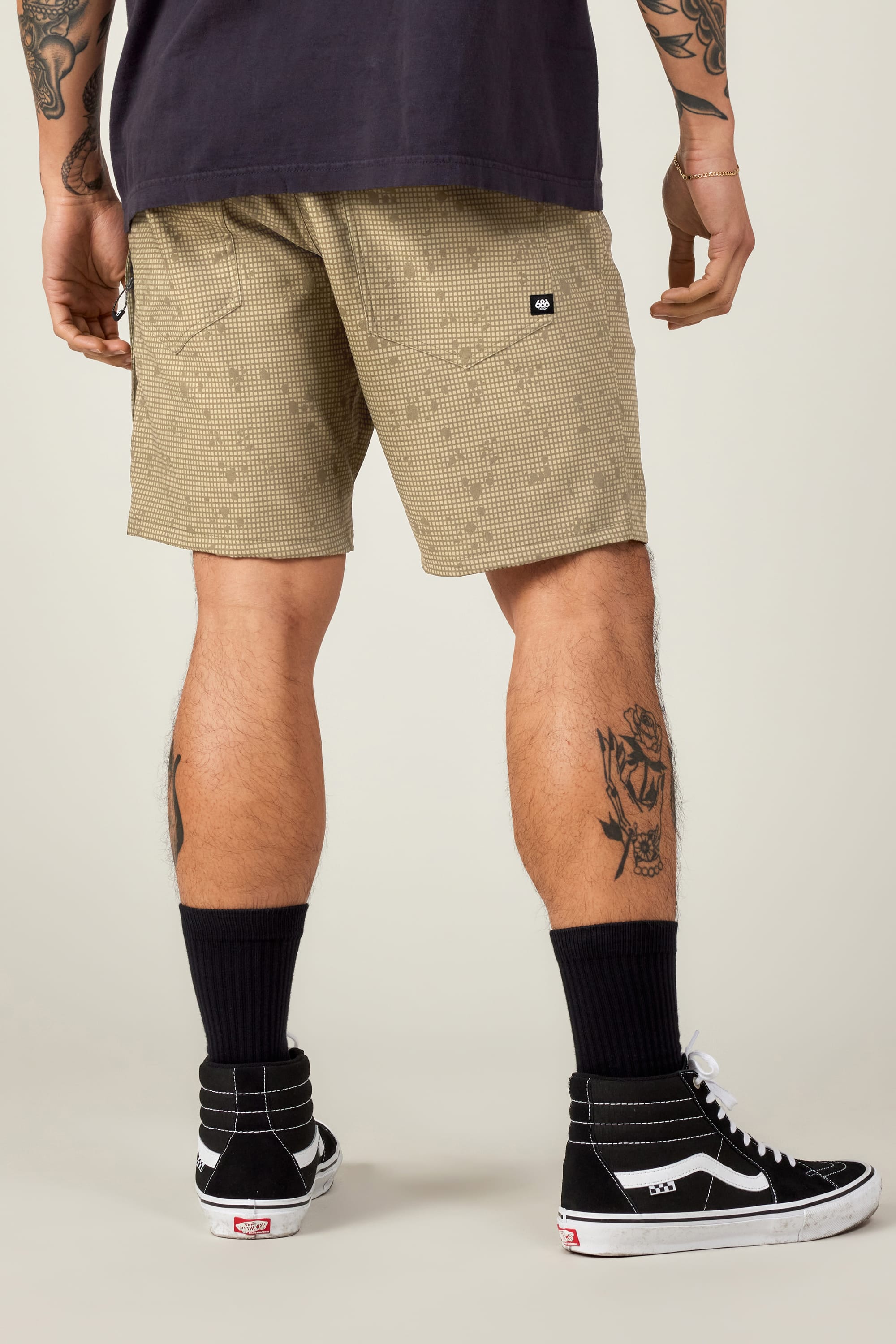 Wearing tan 686 Men's Everywhere® Hybrid Shorts, a black tee, black crew socks, and high-top sneakers, the person stands facing away. The stain-resistant shorts from 686 complement visible arm and leg tattoos.