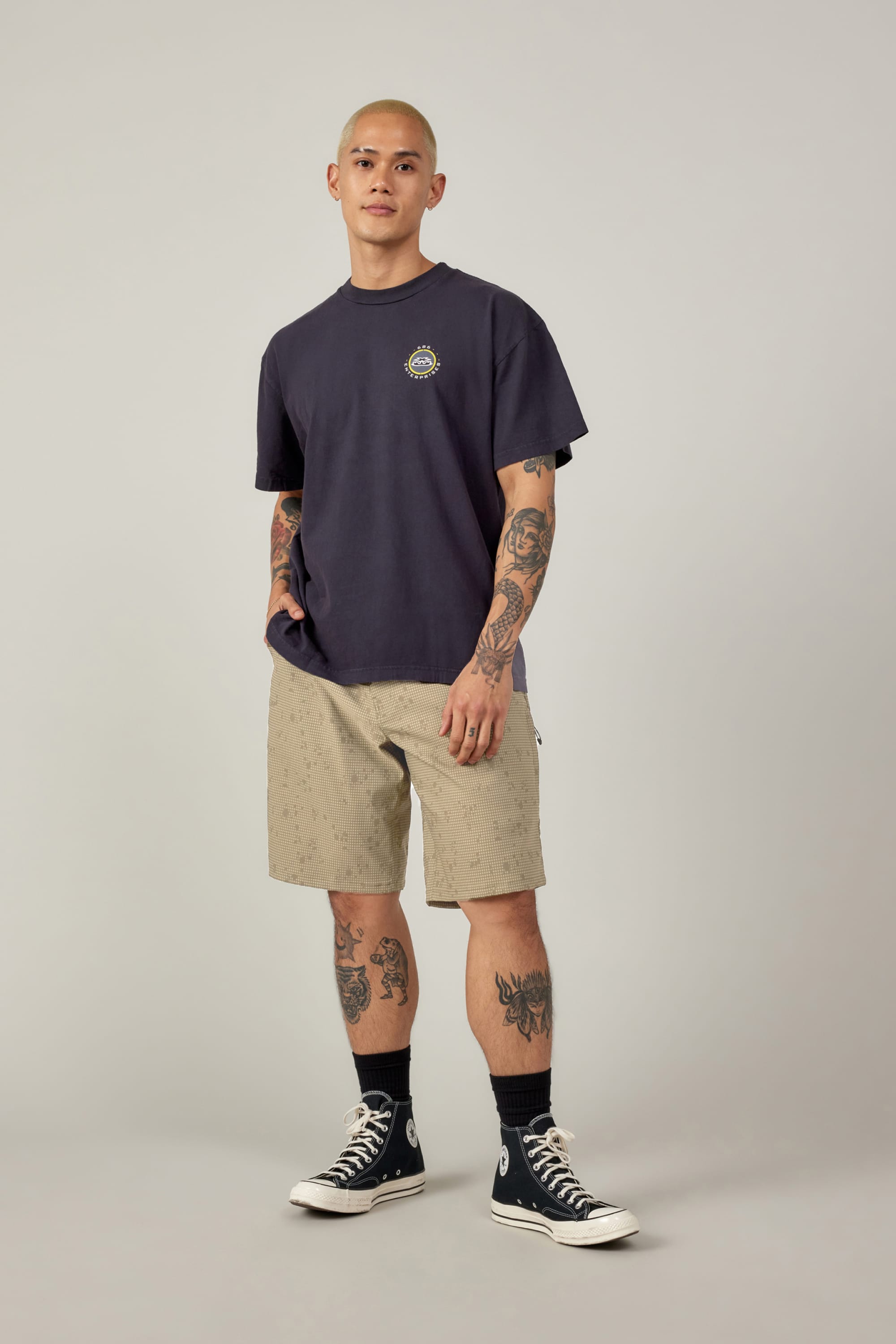 A tattooed person wears a dark navy t-shirt, 686 Men's Everywhere® Hybrid Short - Relaxed Fit in light beige with a subtle pattern, black socks, and black high-top sneakers, standing against a plain light gray background.