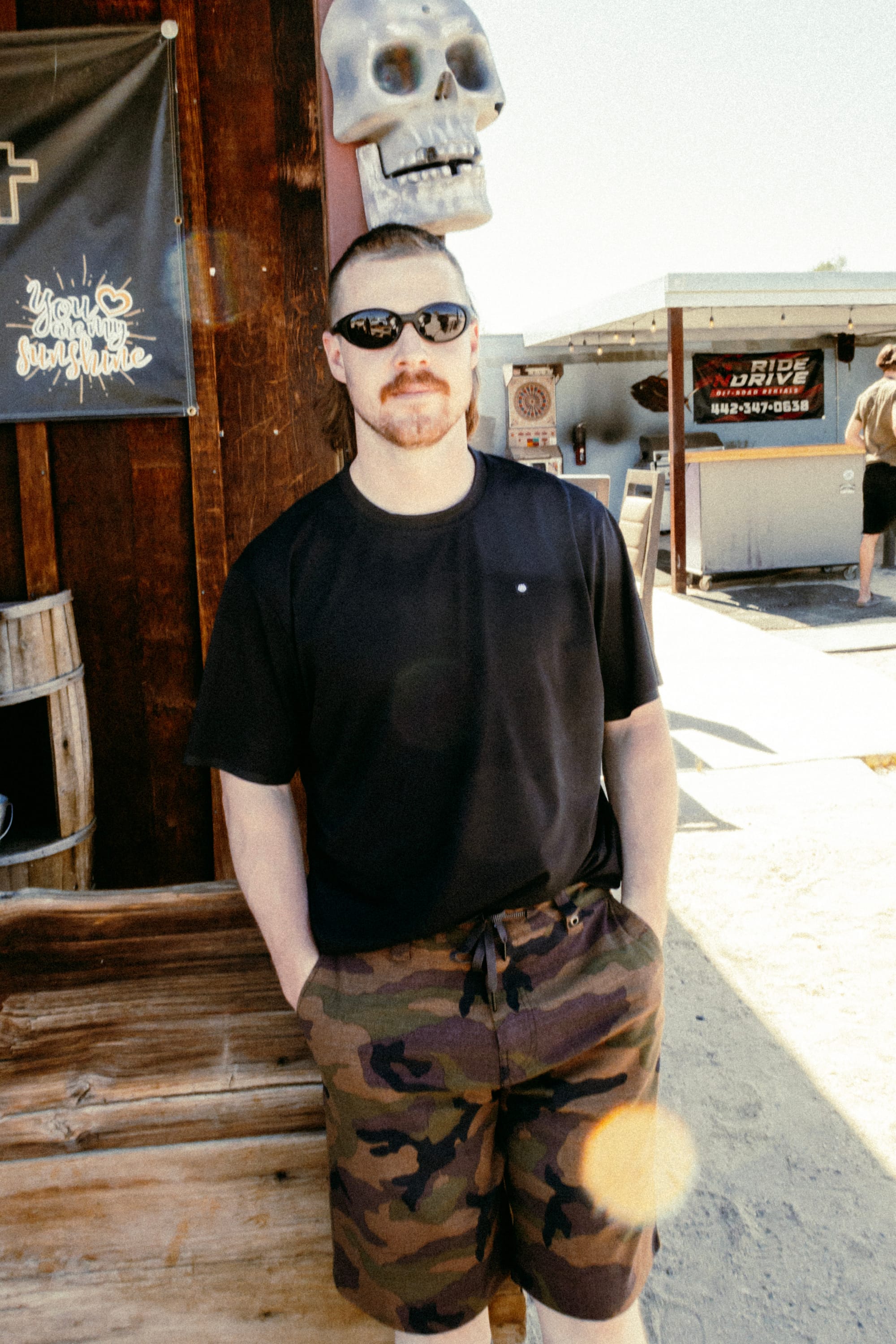A man stands outside in sunlight with his hands in his pockets, wearing a black 686 Mens Lets Go Tech T-Shirt, sunglasses, and camo shorts. A silver skull decoration is attached to the wooden wall above his head.