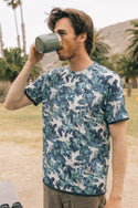 A man with brown hair drinks from a green mug outdoors, wearing the 686 Men's Let's Go Tech Tee by 686—a blue and green leaf-patterned breathable shirt—while standing on grass with palm trees and hills in the background.