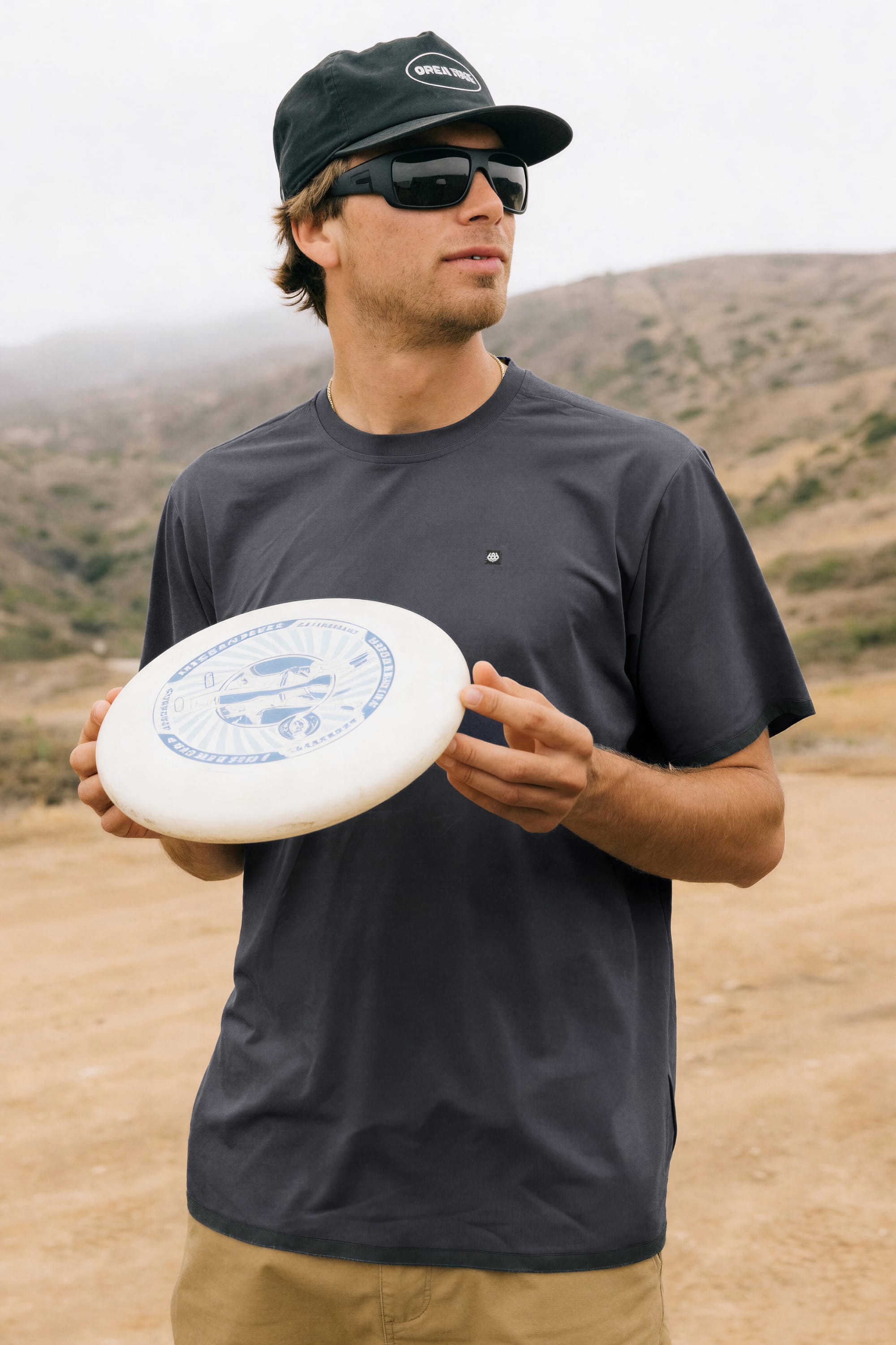 A man in sunglasses and a black cap wears the 686 Men's Let's Go Tech Tee as he holds a white frisbee outdoors, with hills and cloudy skies behind him—ready for spring and summer adventures.