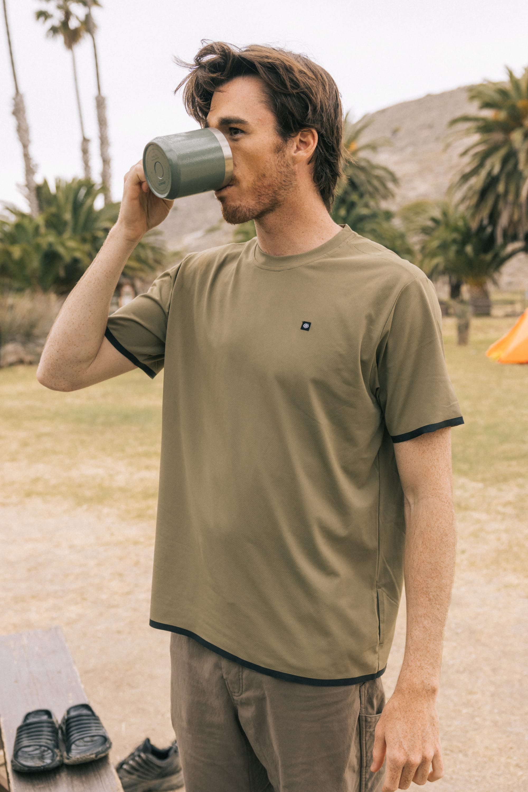 A man in the 686 Mens Lets Go Tech T-Shirt enjoys a drink from a green mug outdoors, surrounded by palm trees, hills, and an orange tent—an ideal scene for spring or summer adventures.