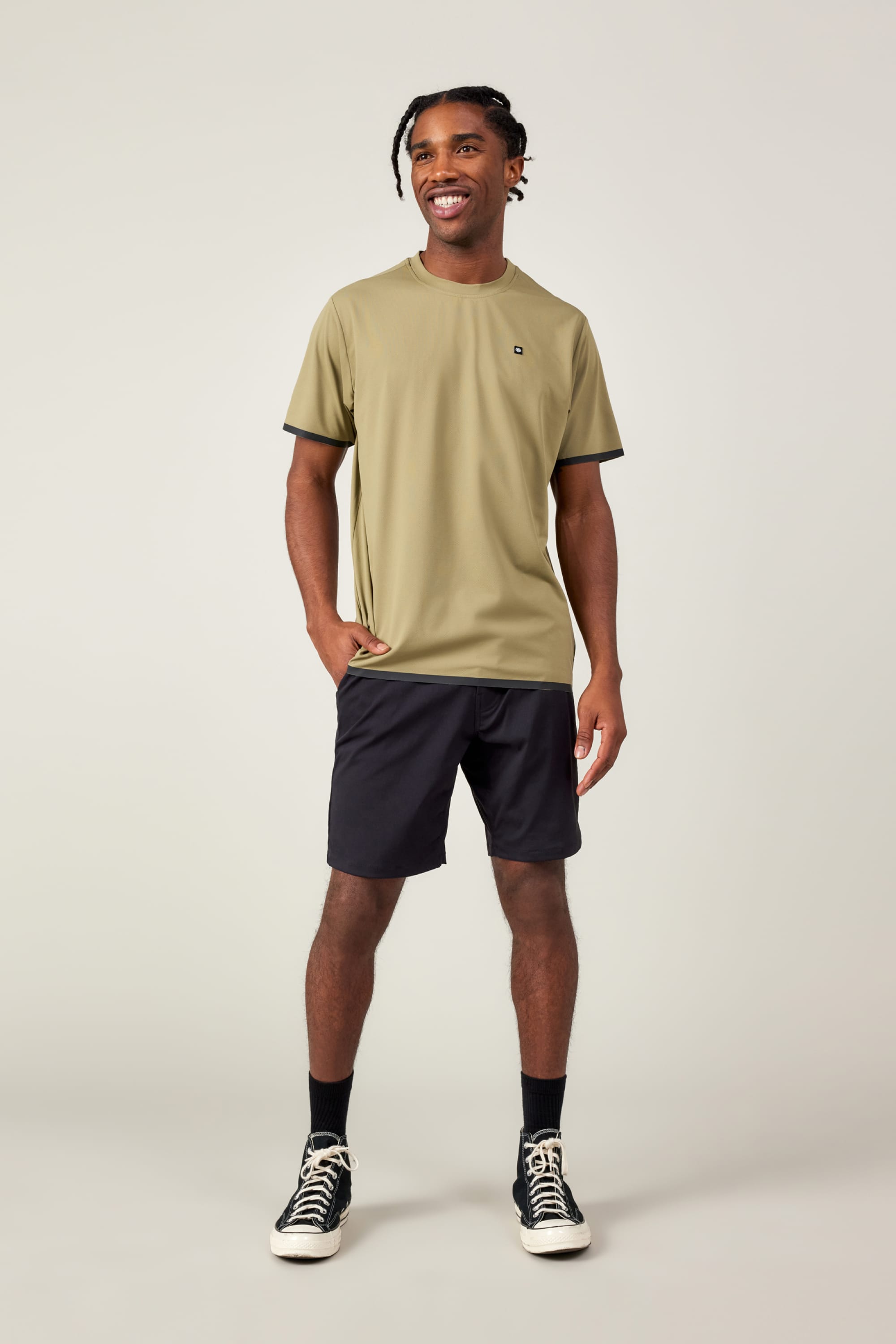 A man smiles against a plain background, wearing the 686 Mens Lets Go Tech T-Shirt by 686 in light olive green, paired with black shorts, black socks, and black sneakers with white soles. One hand is in his pocket; the other hangs by his side.