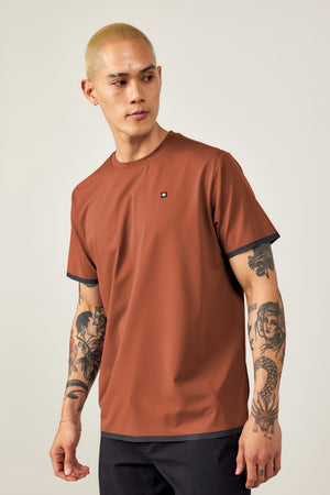 A person with short blond hair and arm tattoos wears the 686 Men's Let's Go Tech Tee in rust, featuring black sleeve trim and paired with black pants, posing against a light background‚Äîideal for spring-summer adventures.