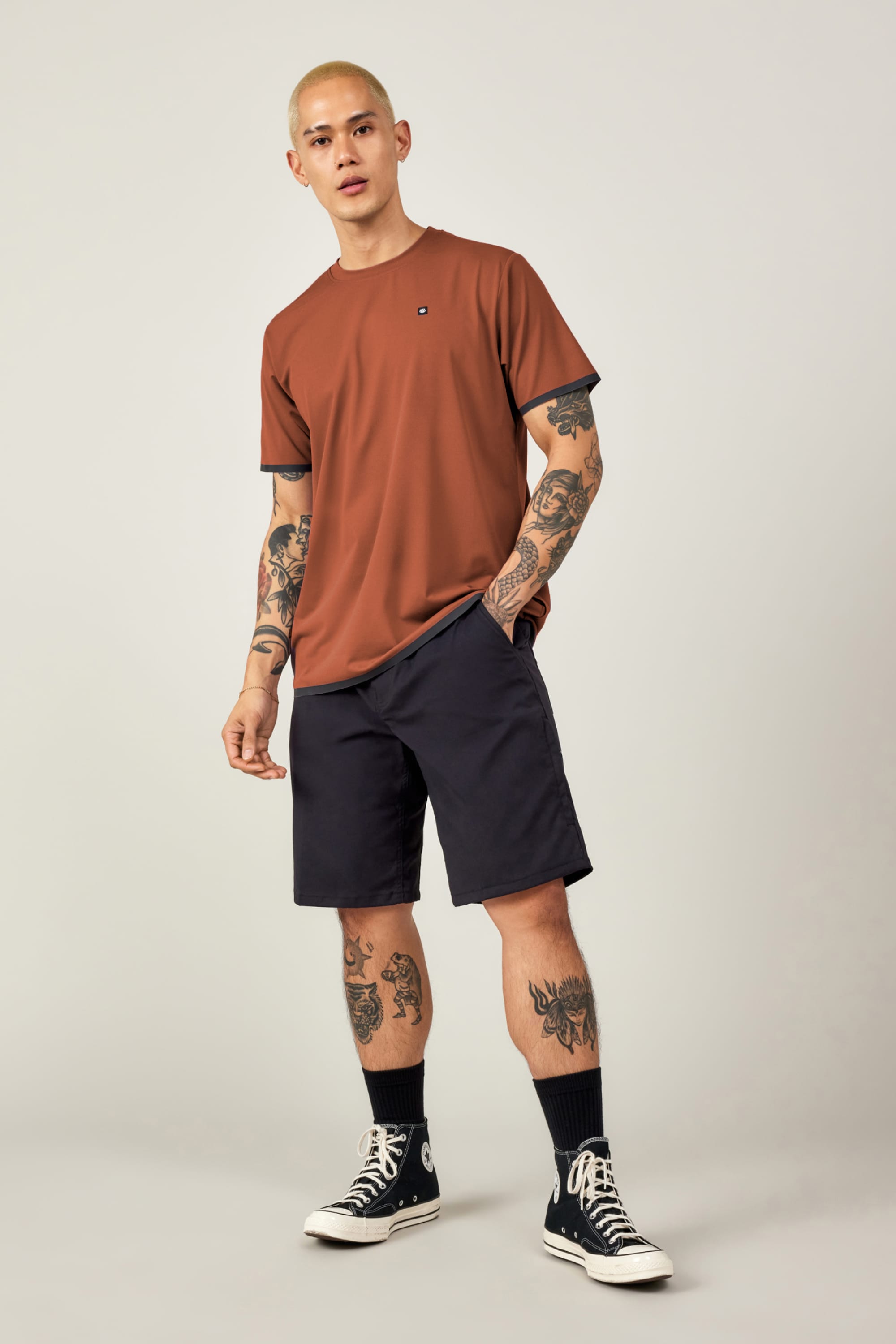 A person with short blond hair and tattoos wears the 686 Men’s Let’s Go Tech Tee by 686—rust-colored, breathable—paired with dark navy shorts, black socks, and high-top sneakers. The perfect outfit for spring-summer adventures against a plain backdrop.