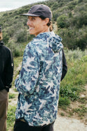 A smiling person wears the 686 Men's Let's Go Tech Hoody with blue and green camo and a dark cap, standing on a dirt path amid greenery and hills, enjoying sun protection in this lightweight, breathable tech piece from 686.