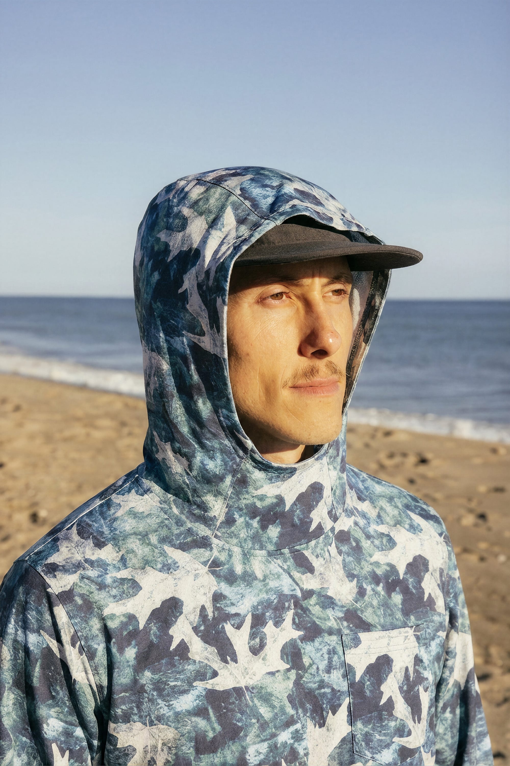 A person in the 686 Men's Let's Go Tech Hoody—a lightweight, blue and white patterned top by 686—stands on a sandy beach, gazing into the distance with a dark cap, ocean waves, and clear sky behind them.