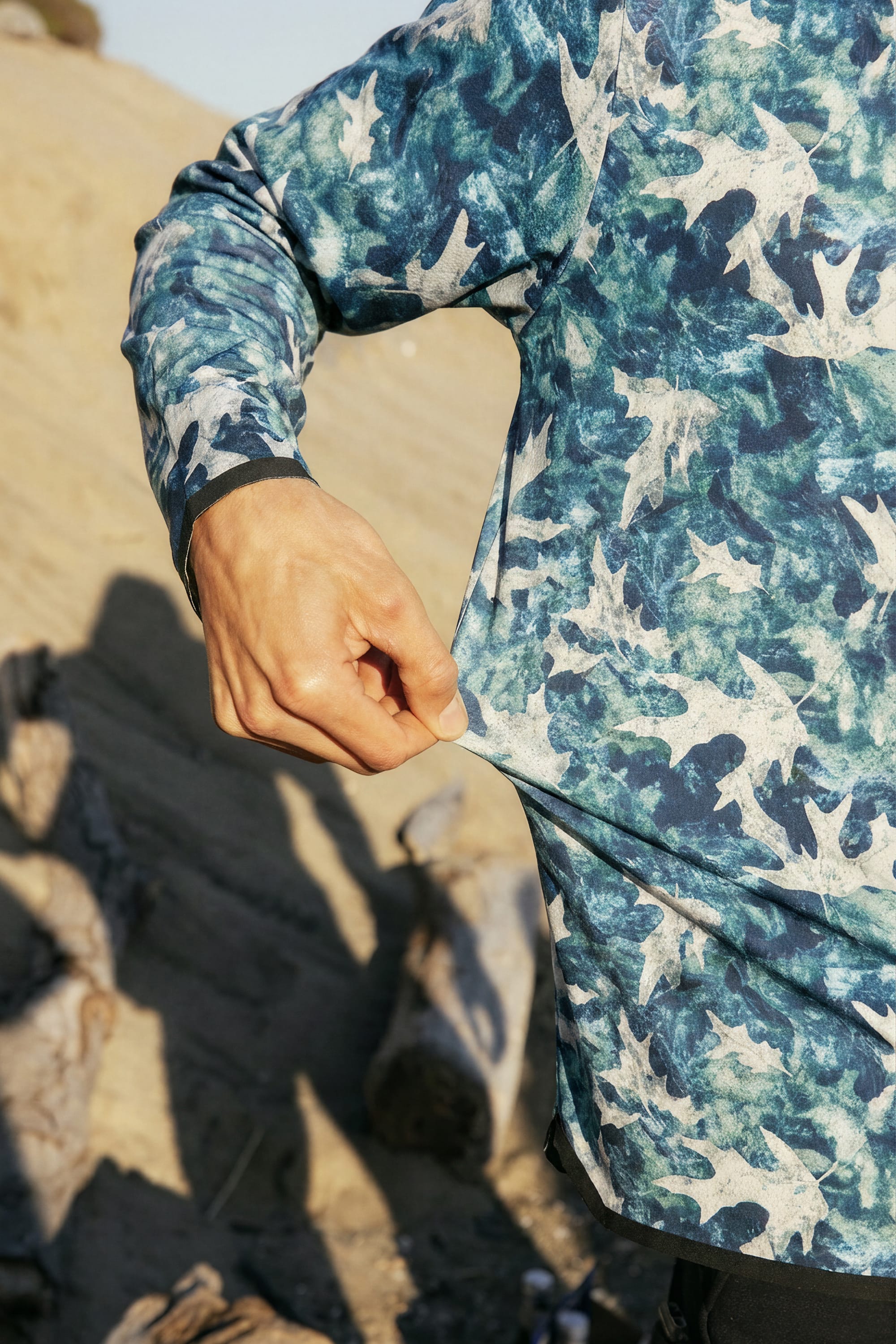 A man wearing the 686 Men's Let's Go Tech Hoody, a lightweight blue-and-white bird-patterned long-sleeve by 686, pulls the fabric at his hip while standing outdoors on sandy terrain with driftwood behind him.