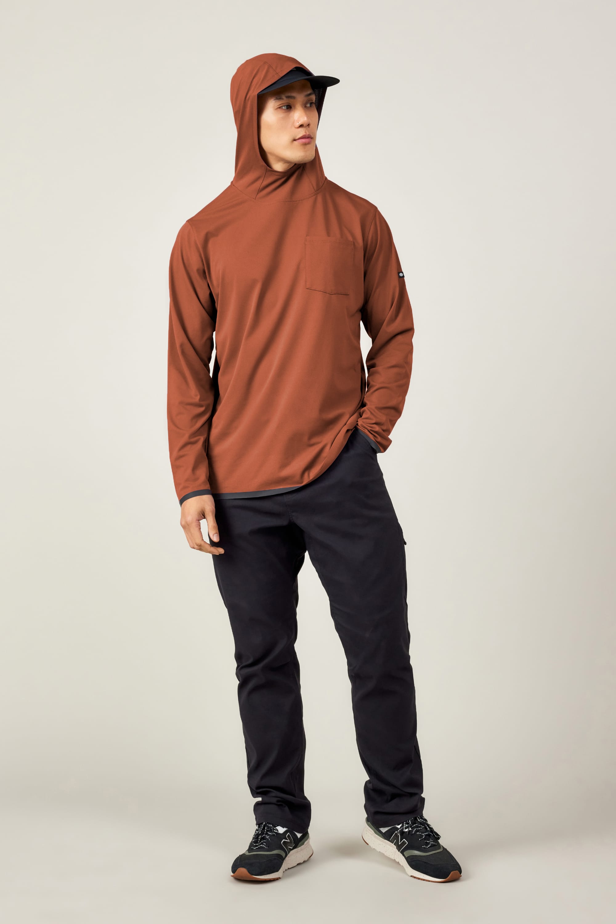 A person stands slightly turned, wearing the 686 Men's Let's Go Tech Hoody in rust for sun protection, paired with black pants and black athletic shoes with white soles, against a plain light background.