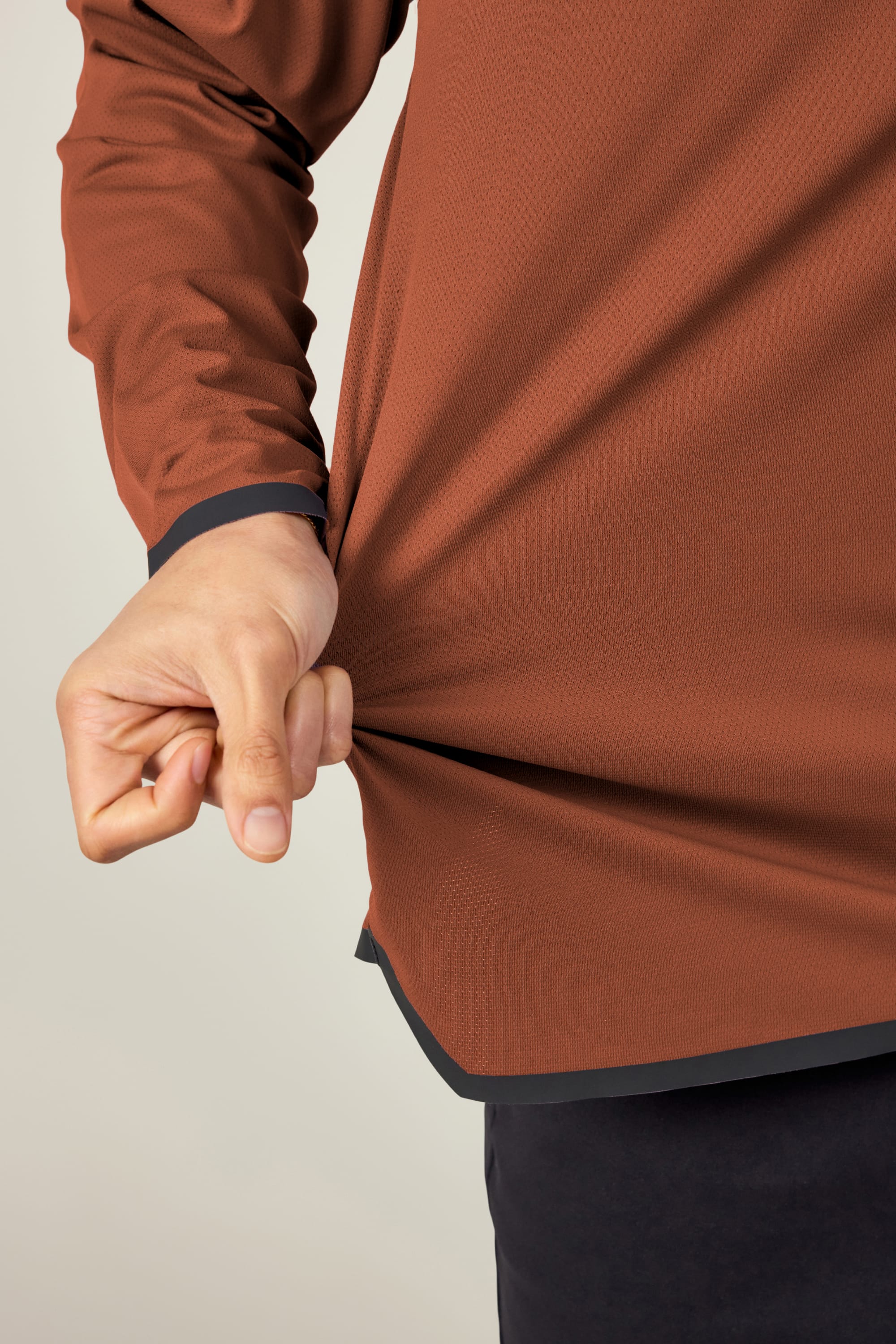 A person wearing the 686 Men's Let's Go Tech Hoody in brown and dark pants pinches and pulls the side of the shirt with their left hand to display its texture and fit. Neutral background. Brand: 686.