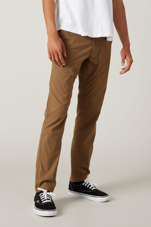 Wearing the 686 Men's Everywhere® Featherlight Chino Pant - Slim Fit in tan, paired with a white t-shirt and black low-top sneakers, a person stands against a light gray background—an ideal summer look. Shown from shoulders to feet.