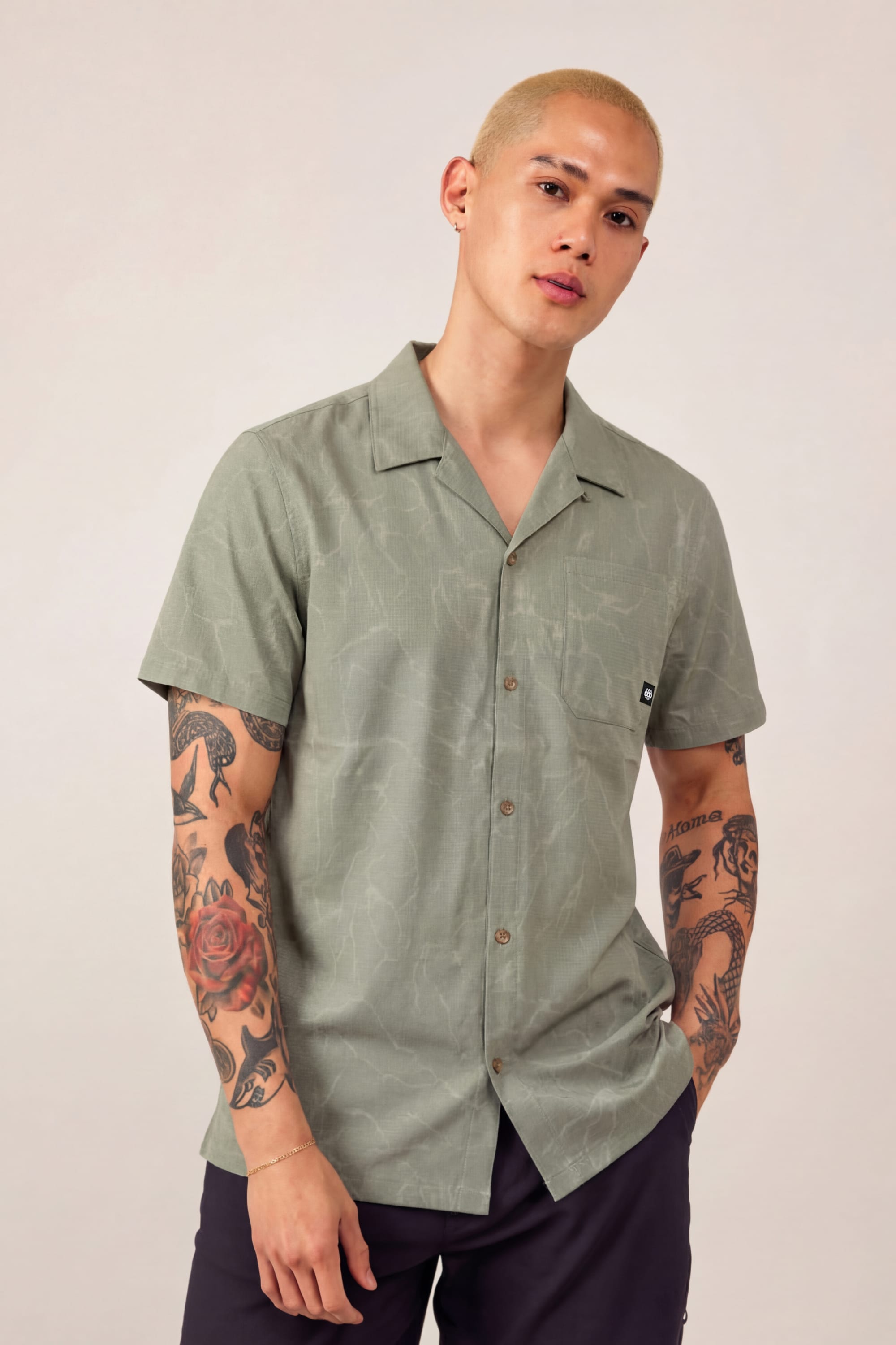 A person with short blond hair and visible arm tattoos models the 686 Men's Nomad Perforated Button Down Shirt in light green, paired with dark pants, standing casually with one hand in their pocket against a plain background.