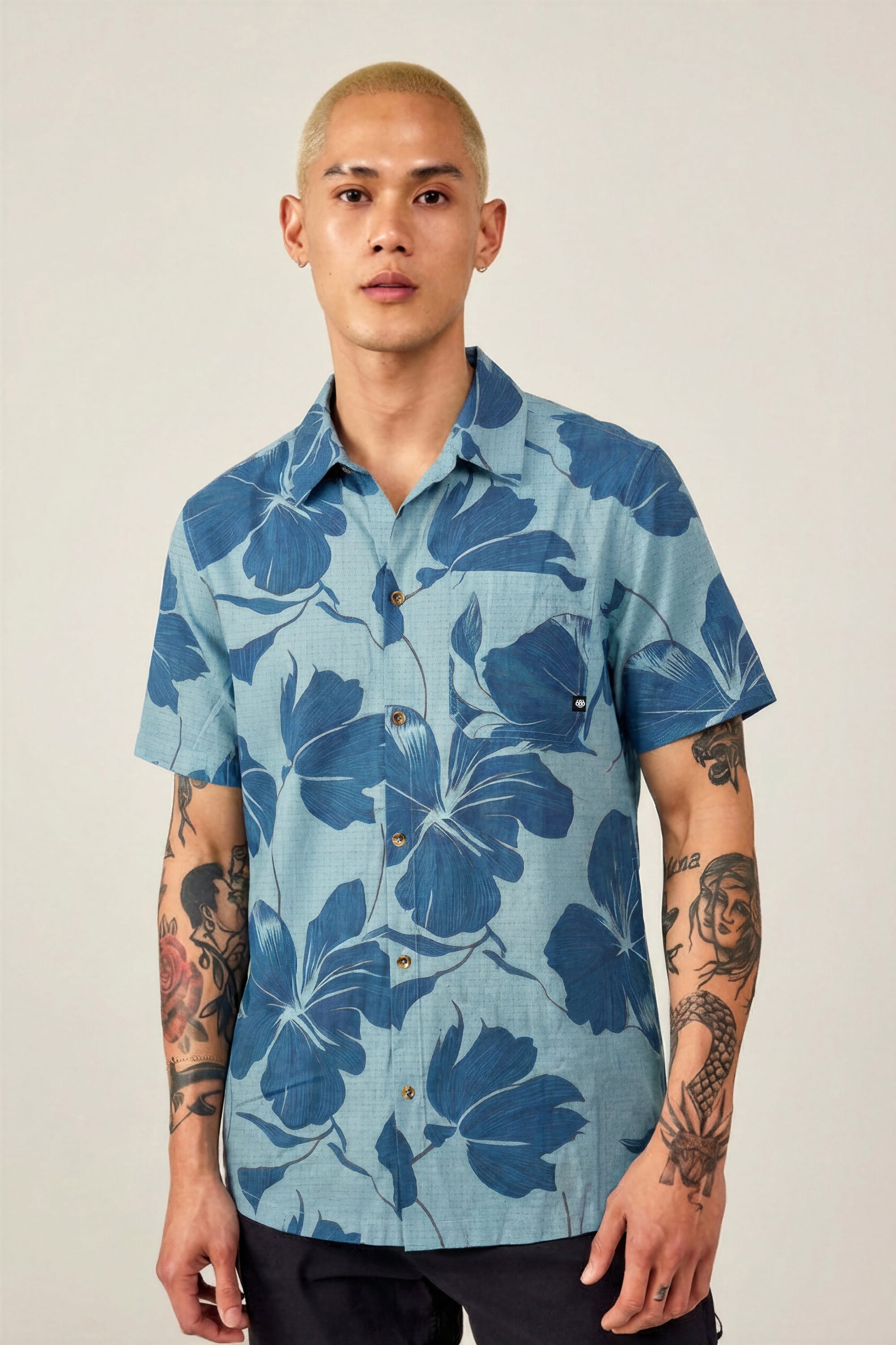 A person with short blond hair and arm tattoos wears the 686 Men's Nomad Perforated Button Down Shirt by 686, featuring a bold dark blue floral pattern, standing facing forward against a plain, light-colored background.