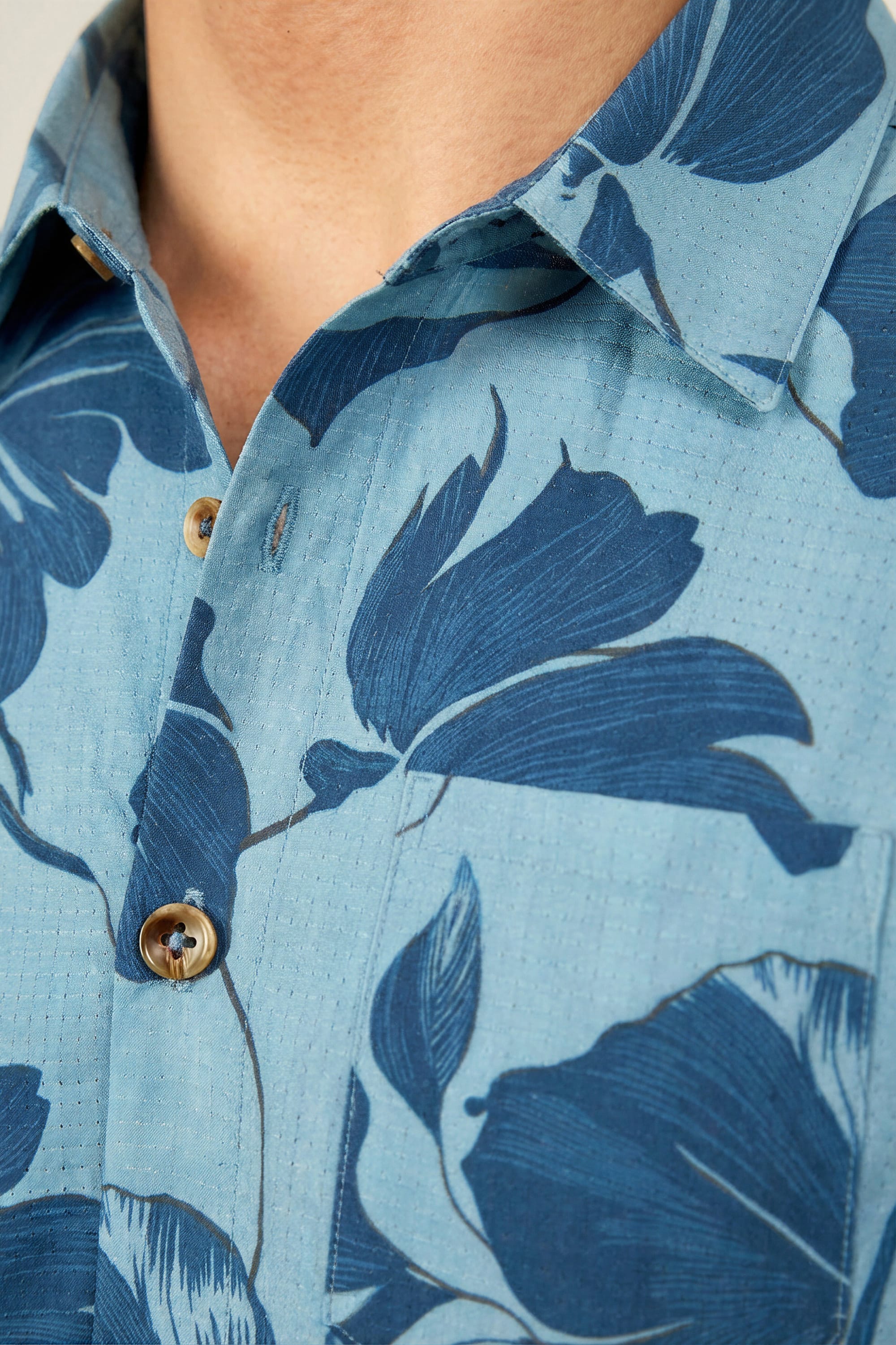 Close-up of a person wearing the 686 Men's Nomad Perforated Button Down Shirt in light blue with dark blue floral patterns and light brown buttons. The image focuses on the upper chest and collar area. Brand: 686.