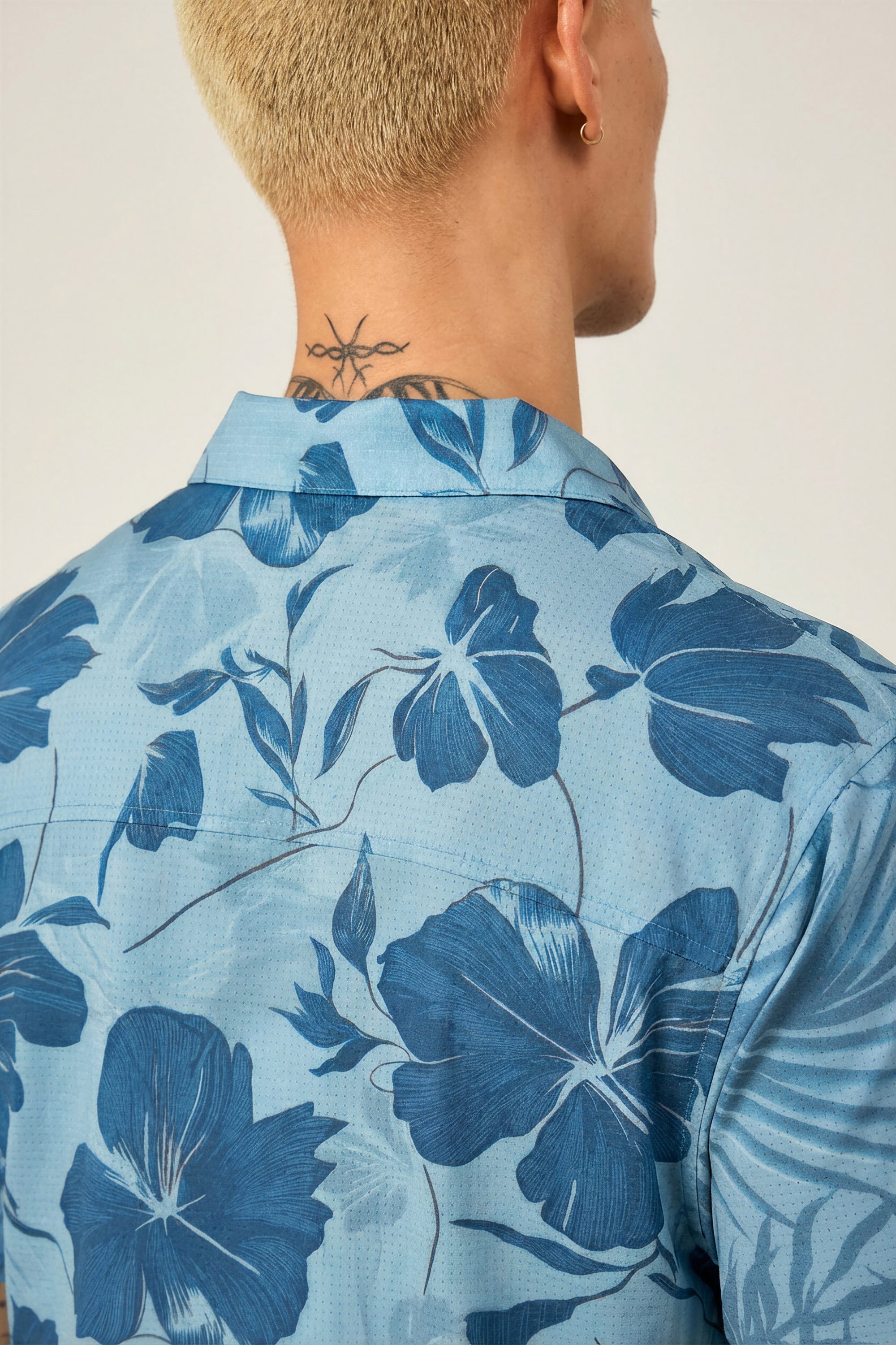 A person with short blond hair and a star tattoo on their neck is seen from behind, wearing the 686 Men's Nomad Perforated Button Down Shirt in light blue with a dark blue floral pattern.