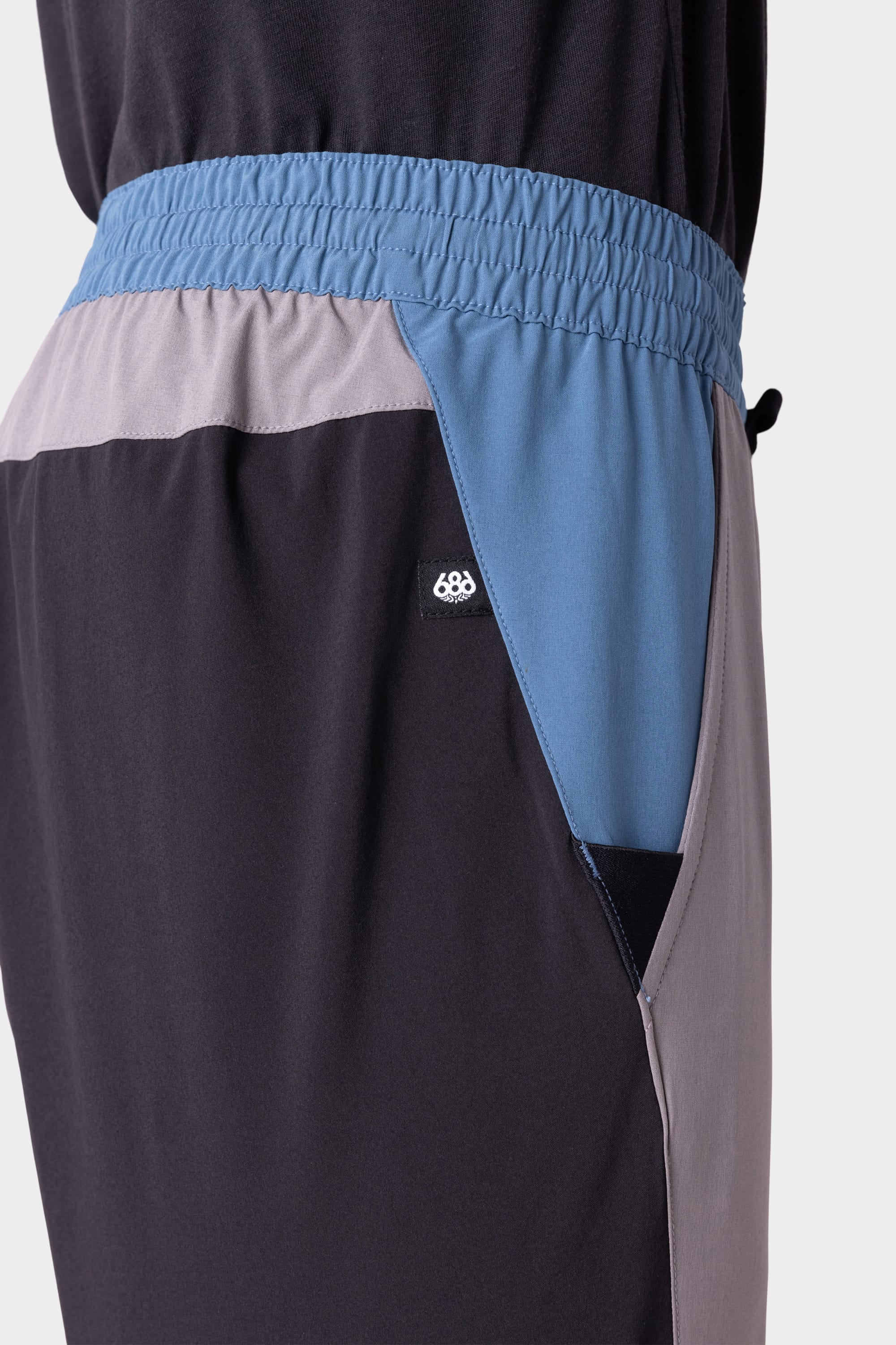 Close-up of the 686 Men's ATP Stretch Performance Pant highlights its black, blue, and gray color-blocked design with an elastic waistband and a small 686 logo tag near the pocket.