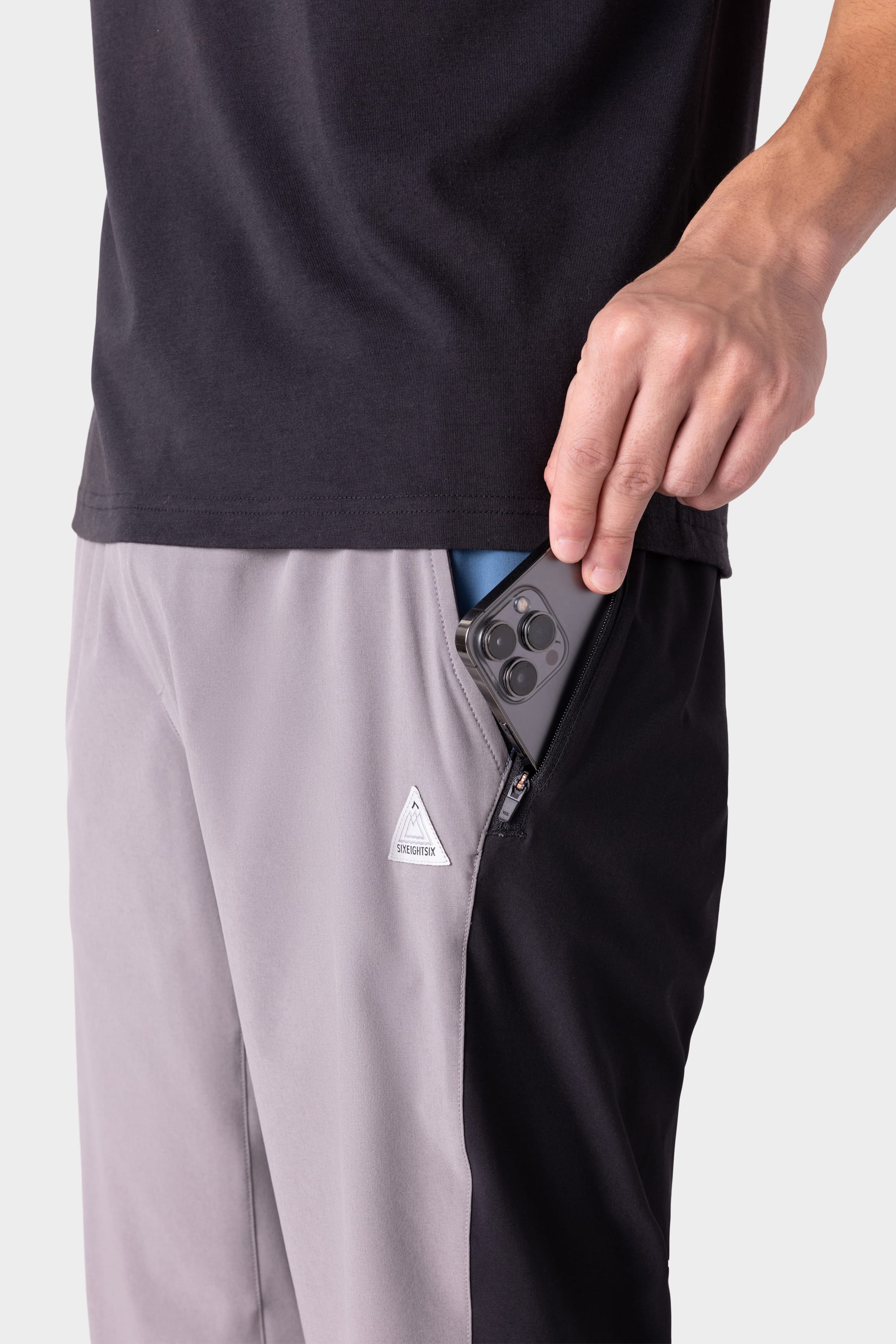 A person wearing 686 Men's ATP Stretch Performance Pant, featuring a triangle logo, slips a smartphone into the zippered side pocket; only their torso and hand are visible.