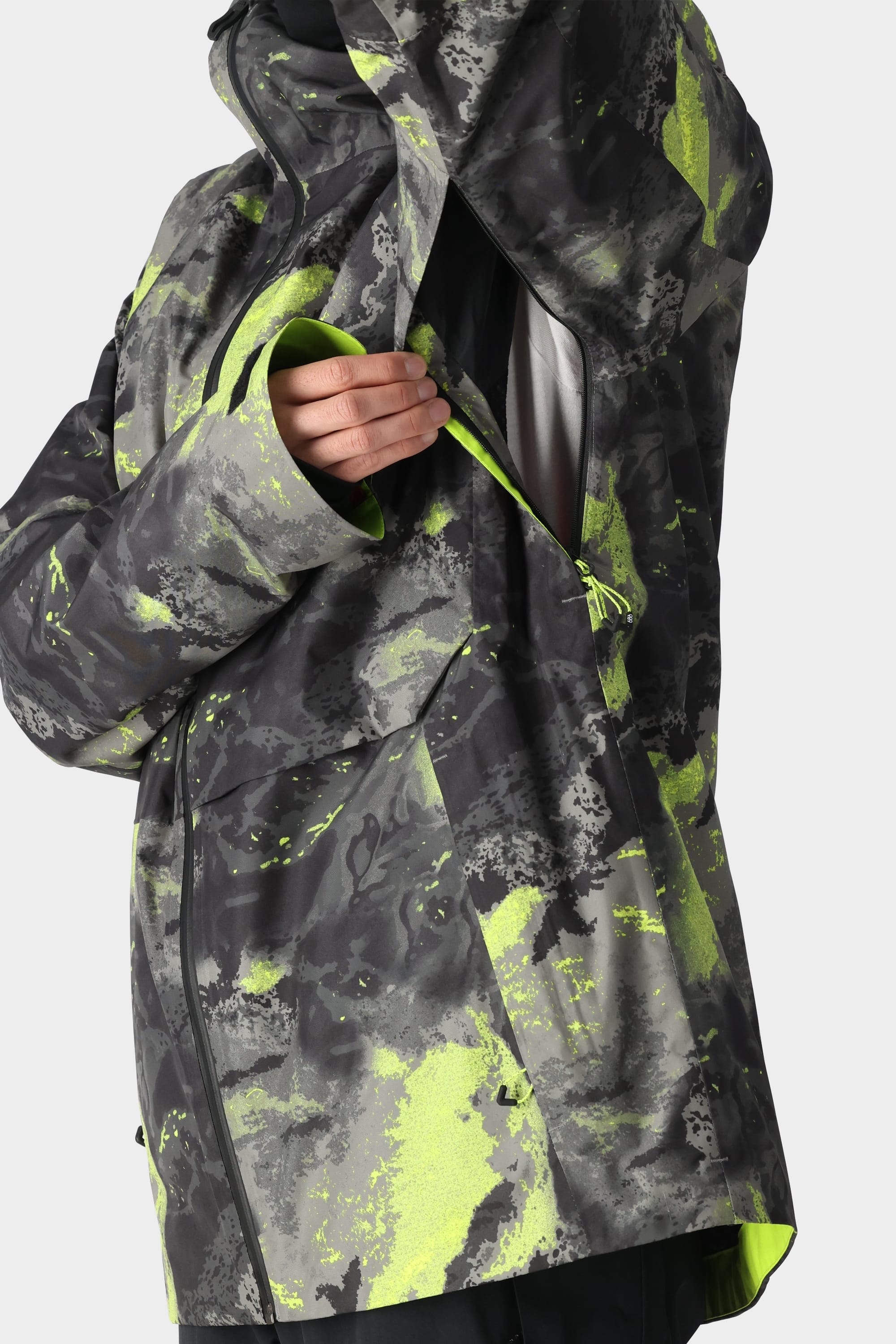 A person wears the 686 Men's GORE-TEX Hydra Down Thermagraph Jacket in black and neon yellow camo, unzipping an underarm vent to show the white body-mapped lining. The background is plain and light-colored.