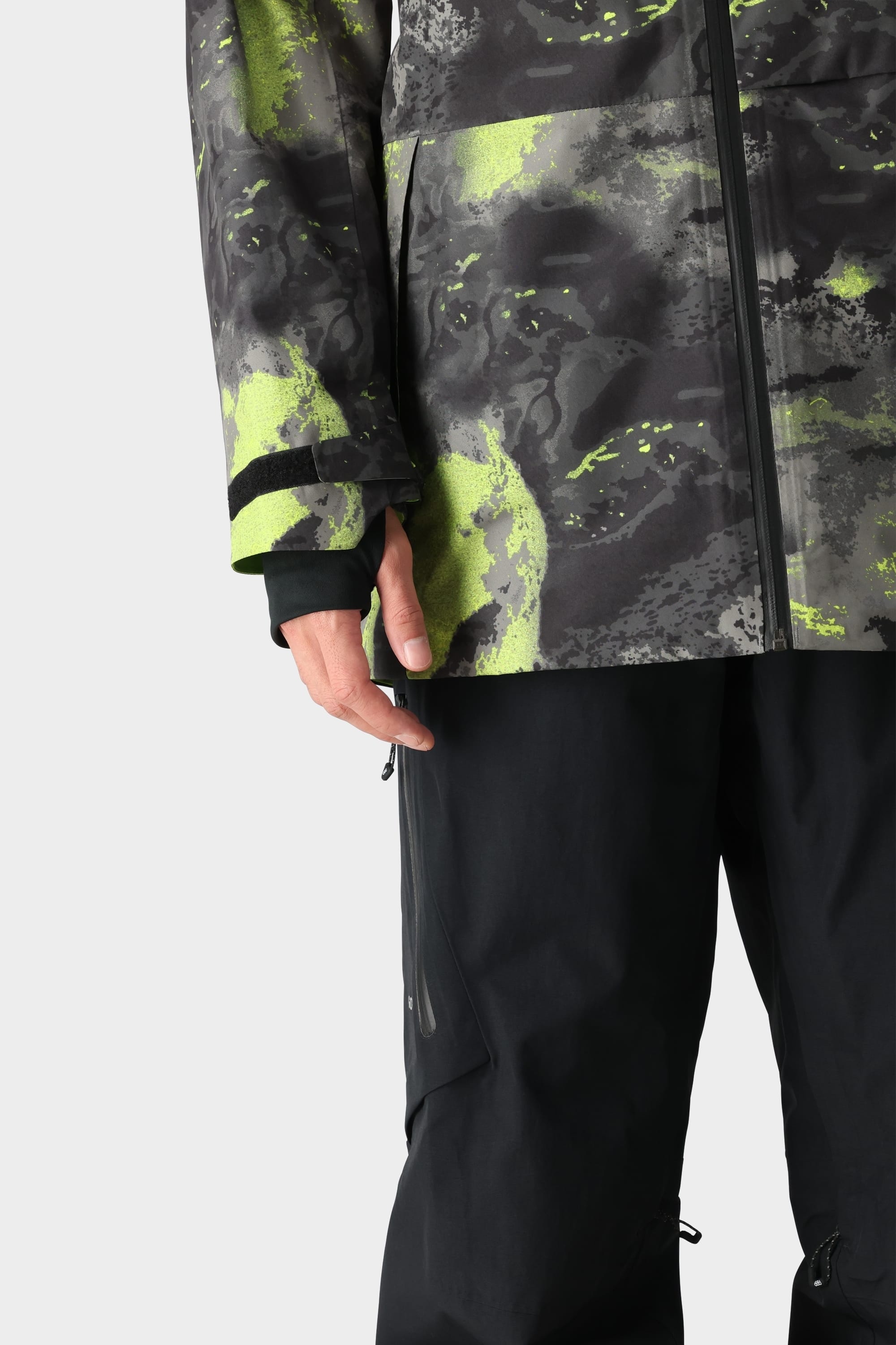 Wearing the 686 Men's GORE-TEX Hydra Down Thermagraph Jacket in black and neon green camo, paired with black pants; only the left hand and lower half are visible against a plain light background.