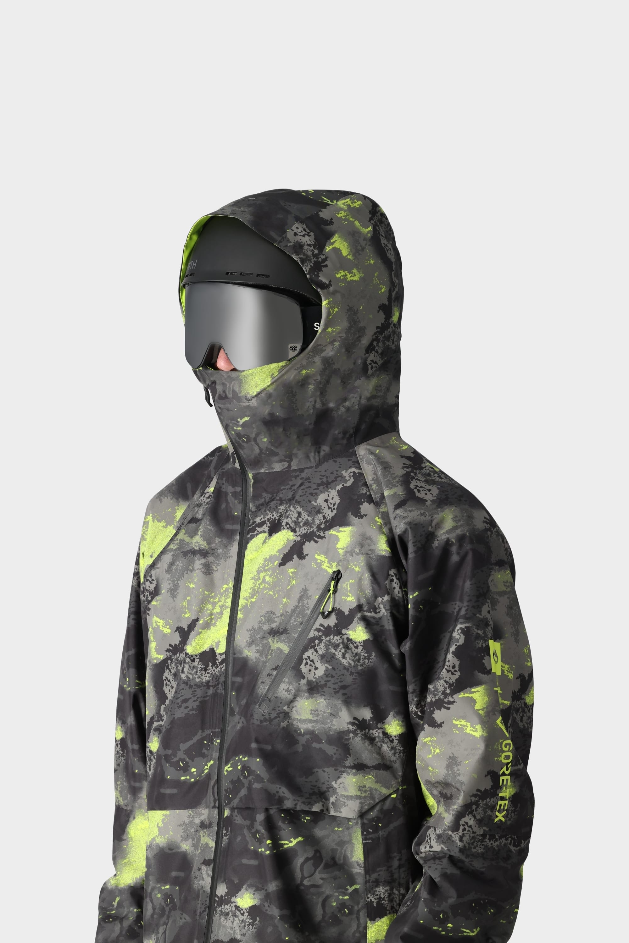 A person wears the 686 Men's GORE-TEX Hydra Down Thermagraph Jacket—a camo hoodie with body-mapped lining, neon yellow-green accents—paired with large reflective goggles, standing against a plain gray background.
