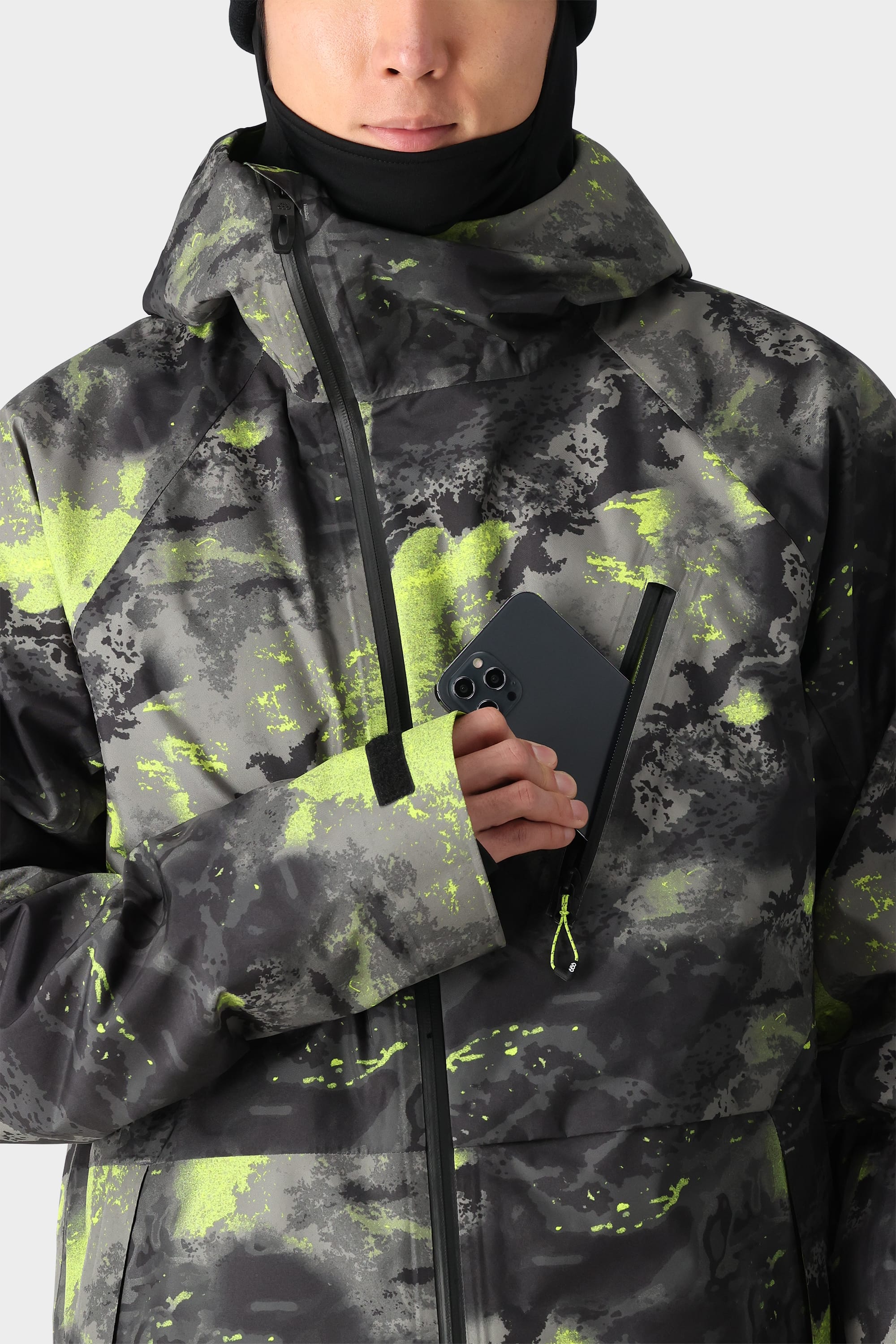A person wearing the 686 Men's GORE-TEX Hydra Down Thermagraph Jacket in black and neon yellow camo, with body-mapped lining, holds a black smartphone partially tucked into the chest zipper pocket. Only their lower face is visible.