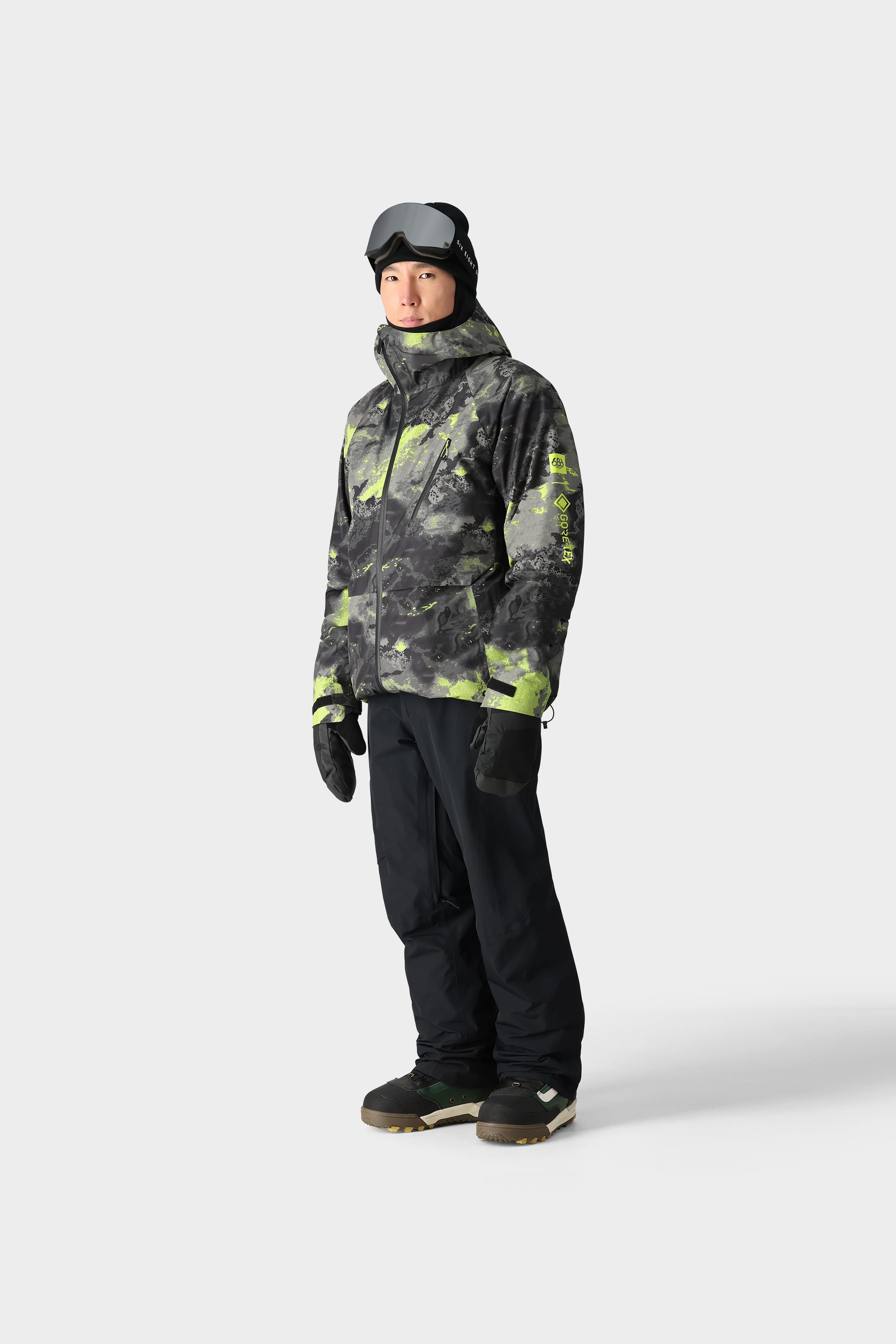 A person in black snow pants, boots, gloves, a black helmet, ski goggles, and a 686 Men's GORE-TEX Hydra Down Thermagraph Jacket stands facing forward against a plain white background.