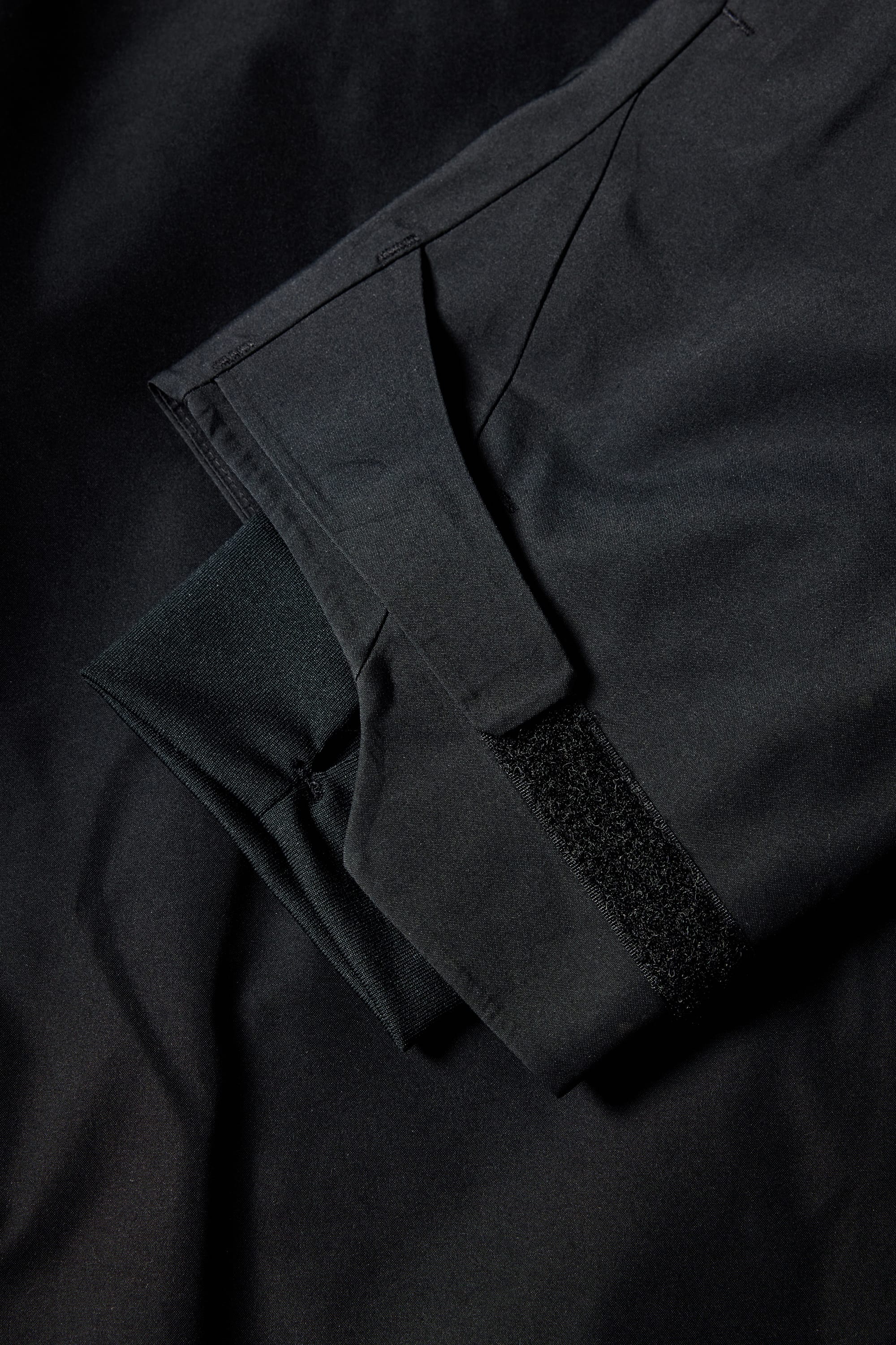 A close-up of the 686 Men's GORE-TEX Hydra Down Thermagraph Jacket shows a Velcro strap and folded fabric with body-mapped lining, highlighting details of this popular black 686 jacket against a dark background.