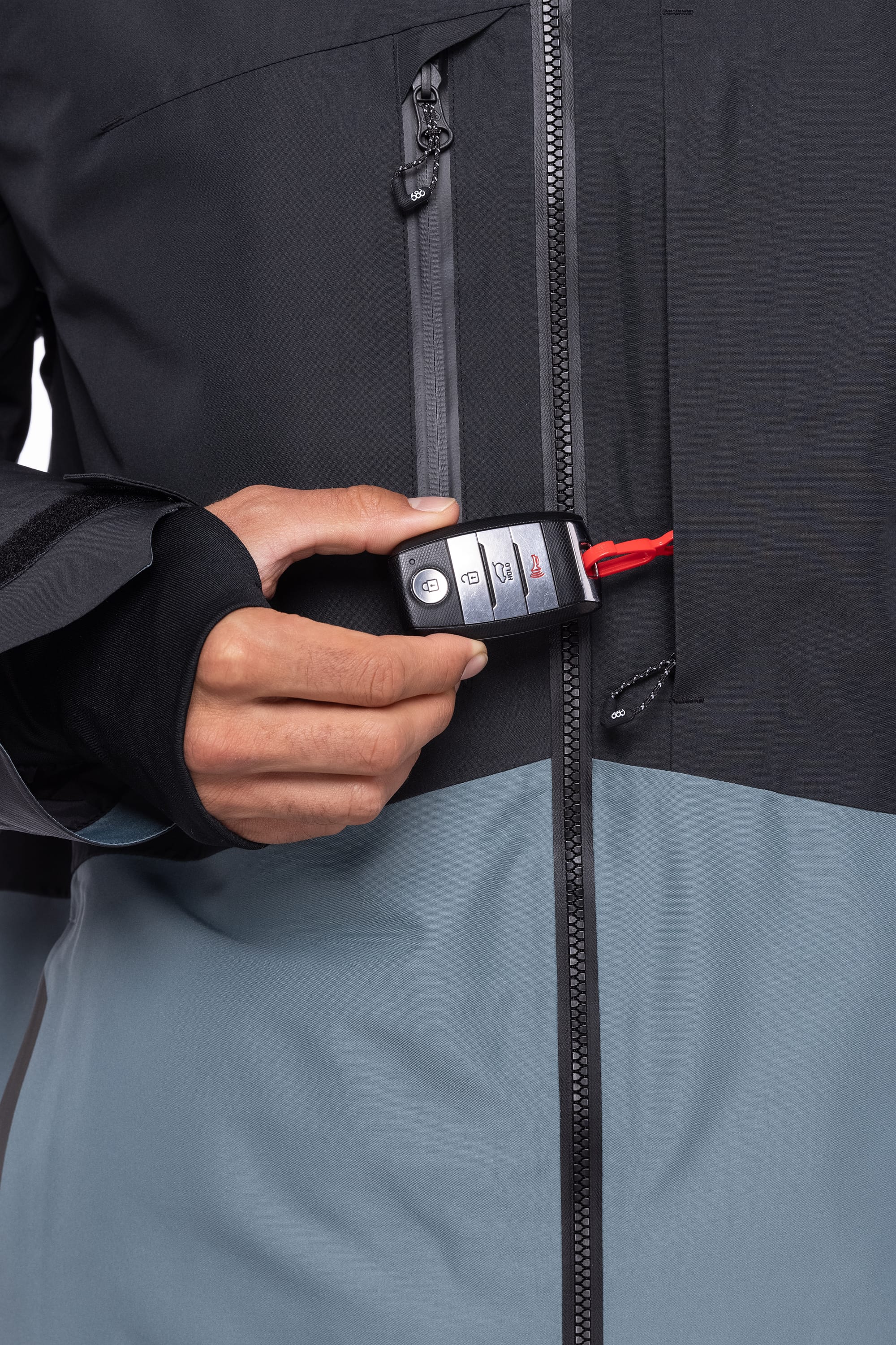 A man wears a 686 Men's Hydrastash Reserve Insulated Jacket in black and gray, holding a car key fob on a red string that's partly tucked into the zippered chest pocket.