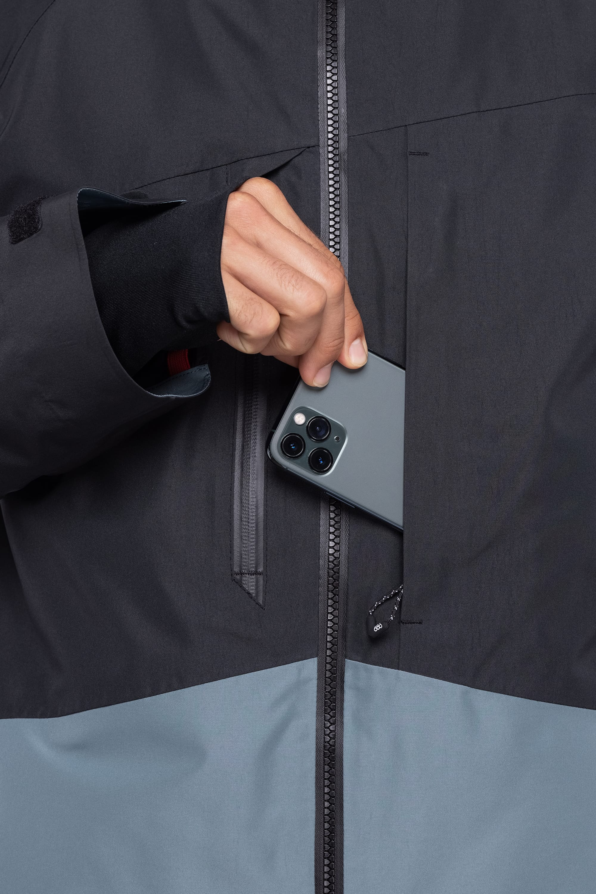 A person wearing the 686 Men's Hydrastash Reserve Insulated Jacket is placing a smartphone with three camera lenses into a zippered chest pocket.