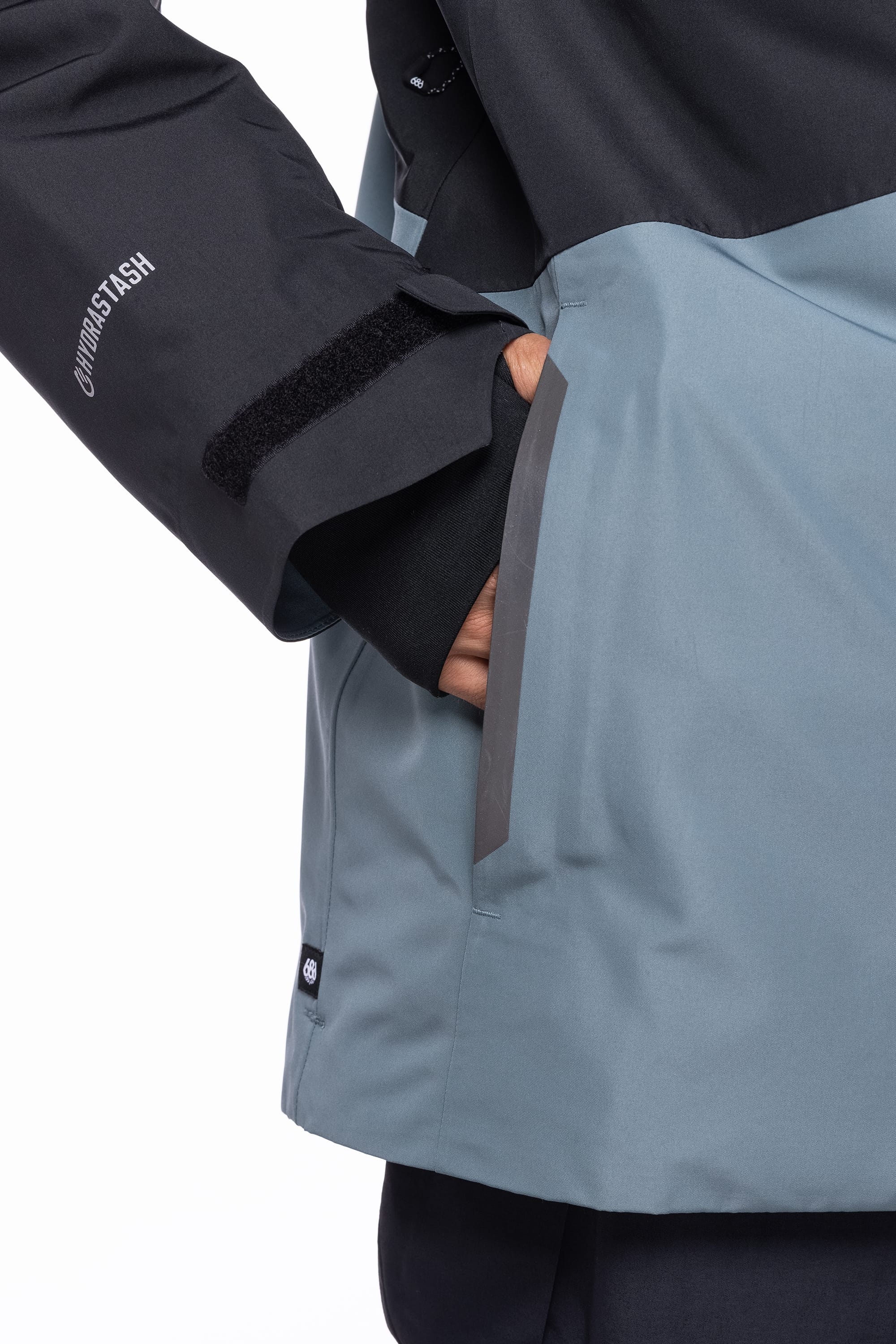 A person wears the 686 Men's Hydrastash Reserve Insulated Jacket in black and gray, featuring a large front pocket, velcro strap on the sleeve, and "UNDERSTASH" printed on the sleeve.